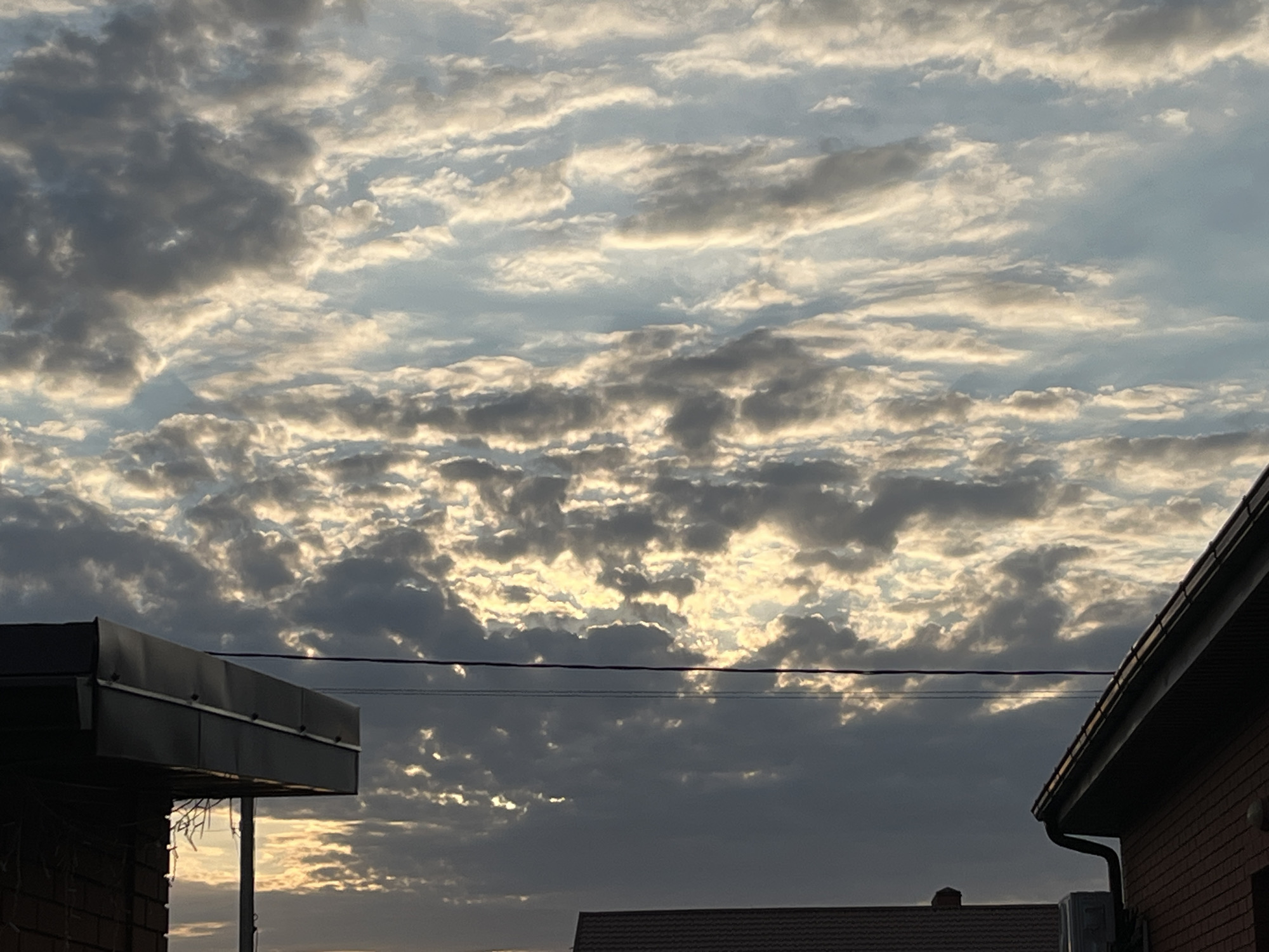 The image shows a cloudy sky with the sun partially obscured by clouds, casting a warm, golden light. In the foreground, the roofs of two buildings are visible on the right and left sides of the image. The building on the left appears to be a single-story structure with a dark, sloped roof and brick walls. The building on the right is a larger, multi-story building with a reddish-brown brick exterior and a dark-colored roof. A power line stretches horizontally across the middle of the image, connecting the two buildings. The scene is set in a residential area, likely in the city of Taganrog City District, Russia, as indicated by the presence of typical urban architecture. The overall impression is one of a serene, late afternoon or early evening sky.