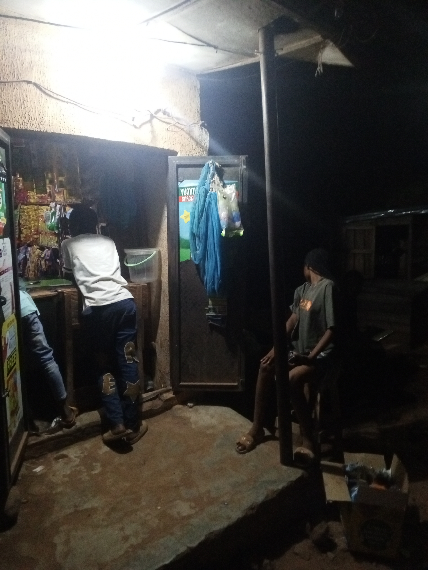 The image depicts an outdoor scene at night, likely in a small urban or rural setting. There is a small, open-front store or kiosk illuminated by a bright light inside. Two people are inside the kiosk: one person with a white shirt is seen interacting with the items on display, and another person is partially visible to the left. Outside the kiosk, to the right, another individual is sitting on a chair or stool. This person appears to be wearing a t-shirt and shorts. A vertical post is near this individual. A cardboard box is on the ground next to the seated individual. A small structure or building is in the background.