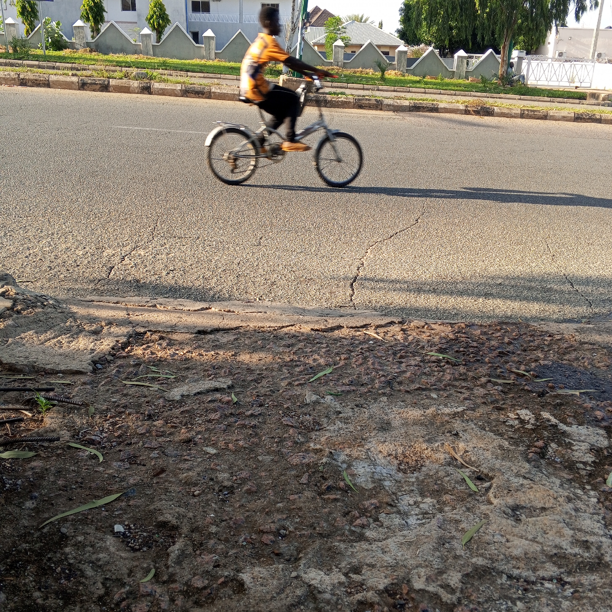 A young boy rides a bicycle on a paved road in Gombe, Nigeria. The boy is wearing an orange and black patterned shirt and dark pants. He is in motion, so his form is blurred. The bicycle is silver with a rear rack. The road appears to be cracked in places and is bordered by a curb. Beyond the curb is a strip of green grass and then a row of buildings with decorative grey facades and peaked roofs. There are also trees and a white gate visible in the background. The sun is low in the sky, casting long shadows across the road, indicating it is likely late afternoon or early morning. The ground in the foreground is rough and uneven, with scattered debris and dry leaves.