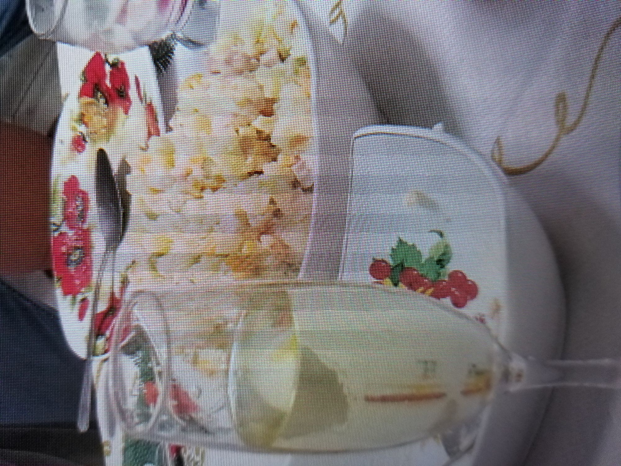The image shows a close-up view of a table setting with food and a drink. In the foreground, a wine glass filled with a light-colored liquid, possibly white wine or champagne, is prominent. The glass is partially obscuring the view of a white serving dish filled with a light-colored, textured food item, likely a salad or rice dish. The dish is decorated with red flowers and green leaves. A fork and knife are placed on the table, suggesting a meal is in progress or about to begin. The background is blurred, but appears to be a domestic setting with dark furniture and a patterned tablecloth. There are no people visible in the image. The image quality is low, characterized by visible pixelation and a lack of sharp detail. The scene is set on a table that is covered with a white tablecloth. There is a hint of a curved, golden decorative element on the tablecloth. The overall impression is that of a casual meal being served.