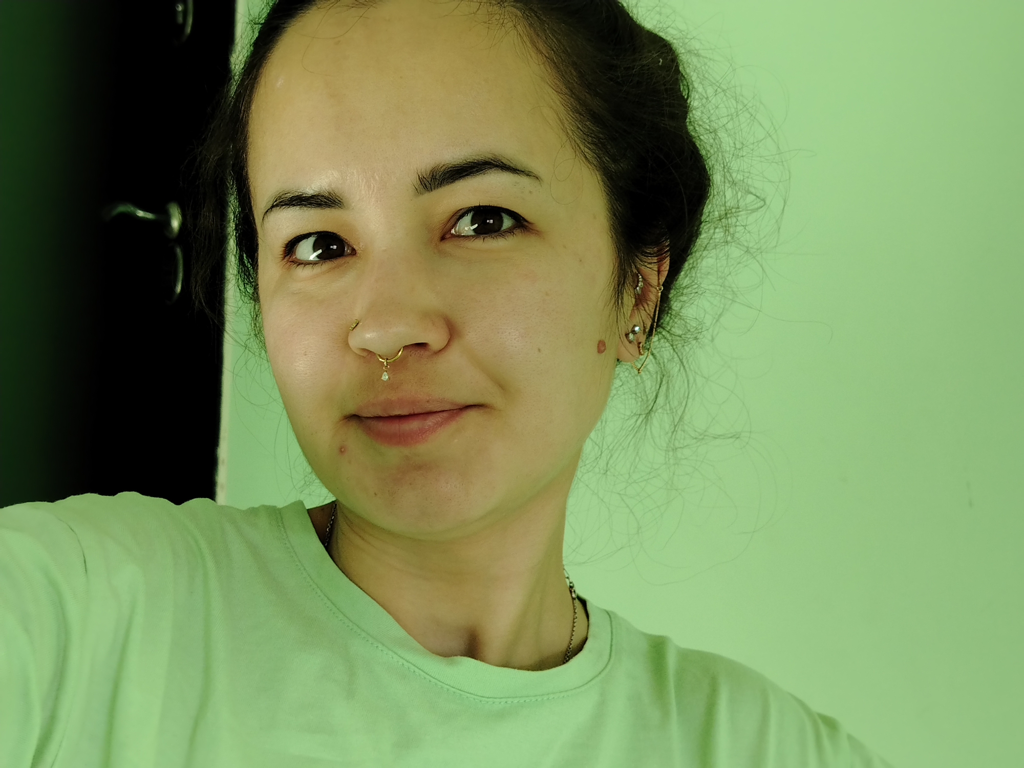 A woman is centered in the frame, wearing a light green t-shirt and a gold nose ring with a pendant. Her dark hair is pulled back, and she has earrings in her left ear. She has a mole on her left cheek. The background is a solid light green wall. A dark door with a silver handle is visible in the upper left corner. The location is unidentifiable as being in Noviy, Russia.