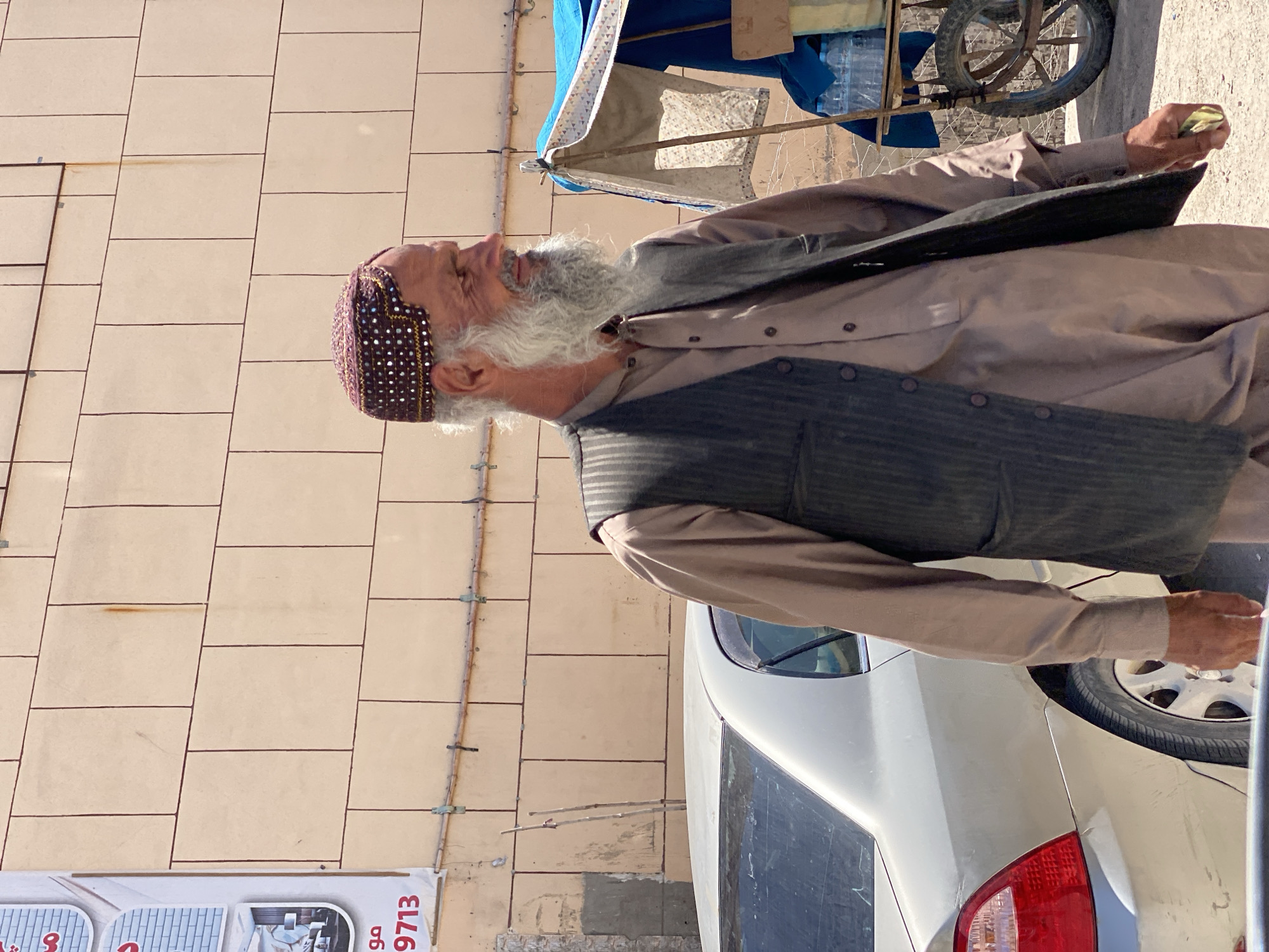 A man with a grey beard and a patterned cap stands on a street in Kandahar, Afghanistan. He wears a brown tunic and a dark vest. A silver sedan is parked behind him. To his right, a cart with a blue tarp is visible, stocked with water bottles. The background features a light-colored building with a grid pattern of darker lines.