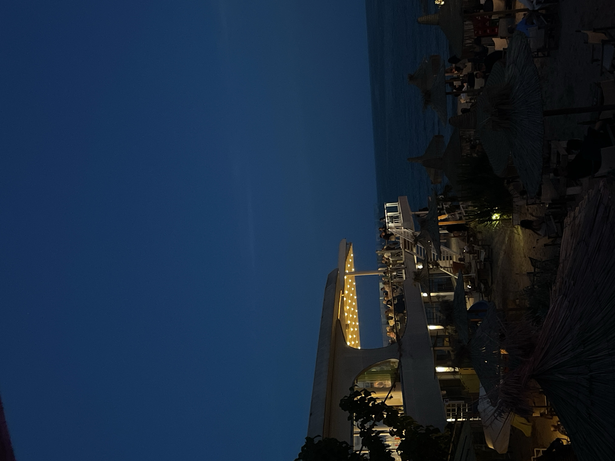 This nighttime image captures a multi-level beachside restaurant and bar located in Varna, Bulgaria. The upper third of the frame shows a deep blue, clear night sky. Below, the dark expanse of the Black Sea occupies the middle-right and background, meeting the horizon line.
The central structure is a modern, light-colored building featuring a distinctive curved roofline and large windows. Its prominent upper terrace is extensively illuminated by multiple strands of warm, downward-facing string lights. Numerous individuals are seated at tables on this brightly lit terrace, engaged in social activity. Below this main terrace, additional architectural elements and staircases are visible, connecting various levels of the establishment.
The sandy beach area, extending from the foreground to the right, is furnished with multiple large, conical thatched umbrellas. Underneath these umbrellas and surrounding them, tables and chairs are positioned, many of which are occupied by patrons. The beach level is significantly darker than the illuminated terrace, relying on ambient light sources. Dark, leafy foliage partially obscures the left side of the building in the foreground. No specific actions beyond sitting and socializing are discernible among the numerous individuals present.