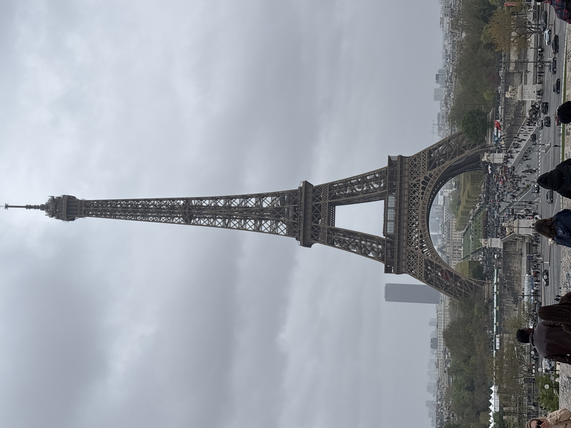 The image shows the Eiffel Tower in Paris, France, on a cloudy day. The tower is made of metal and has a distinctive latticework structure. It stands tall against the gray sky. At the base of the tower, there is a large crowd of people, suggesting it is a popular tourist destination. A road with cars and other traffic runs alongside the base of the tower. Trees and other buildings are visible in the background, indicating the tower's central location within the city. The angle suggests the photo was taken from an elevated position, likely a nearby viewing platform or hill.