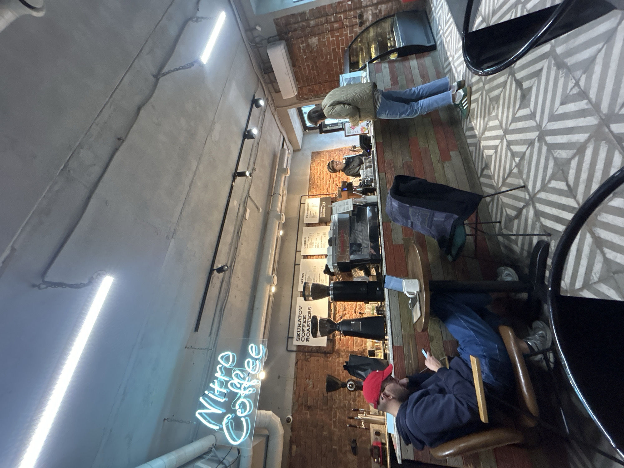 Here is a forensic description of the image.
**Location:** The image appears to be taken inside a coffee shop called "Nitro Coffee."
**Environment:** The interior of the coffee shop has a mix of exposed brick walls, a concrete ceiling with exposed pipes and lighting, and modern furniture. The floor is tiled with a geometric pattern.
**People:**
* One person is standing at the counter, possibly placing an order or waiting to be served.
* Another person is seated at a table, looking at their mobile phone. They are wearing a red cap and a dark-colored jacket.
* There are also other people near the counter who seem to be employees.
**Objects:**
* A neon sign displays the name "Nitro Coffee."
* The counter is equipped with coffee brewing equipment.
* Menu boards with information are visible behind the counter.
* The furniture includes chairs, tables, and possibly bar stools.
* Mobile phone.
**Actions:**
* One person is browsing or using their mobile phone.
* Another person seems to be ordering or being served at the counter.
* The employees appear to be working, possibly preparing drinks or serving customers.
**Interactions:**
* There might be an interaction between the customers and the employees at the counter.
* The person seated at the table is engaged with their phone.