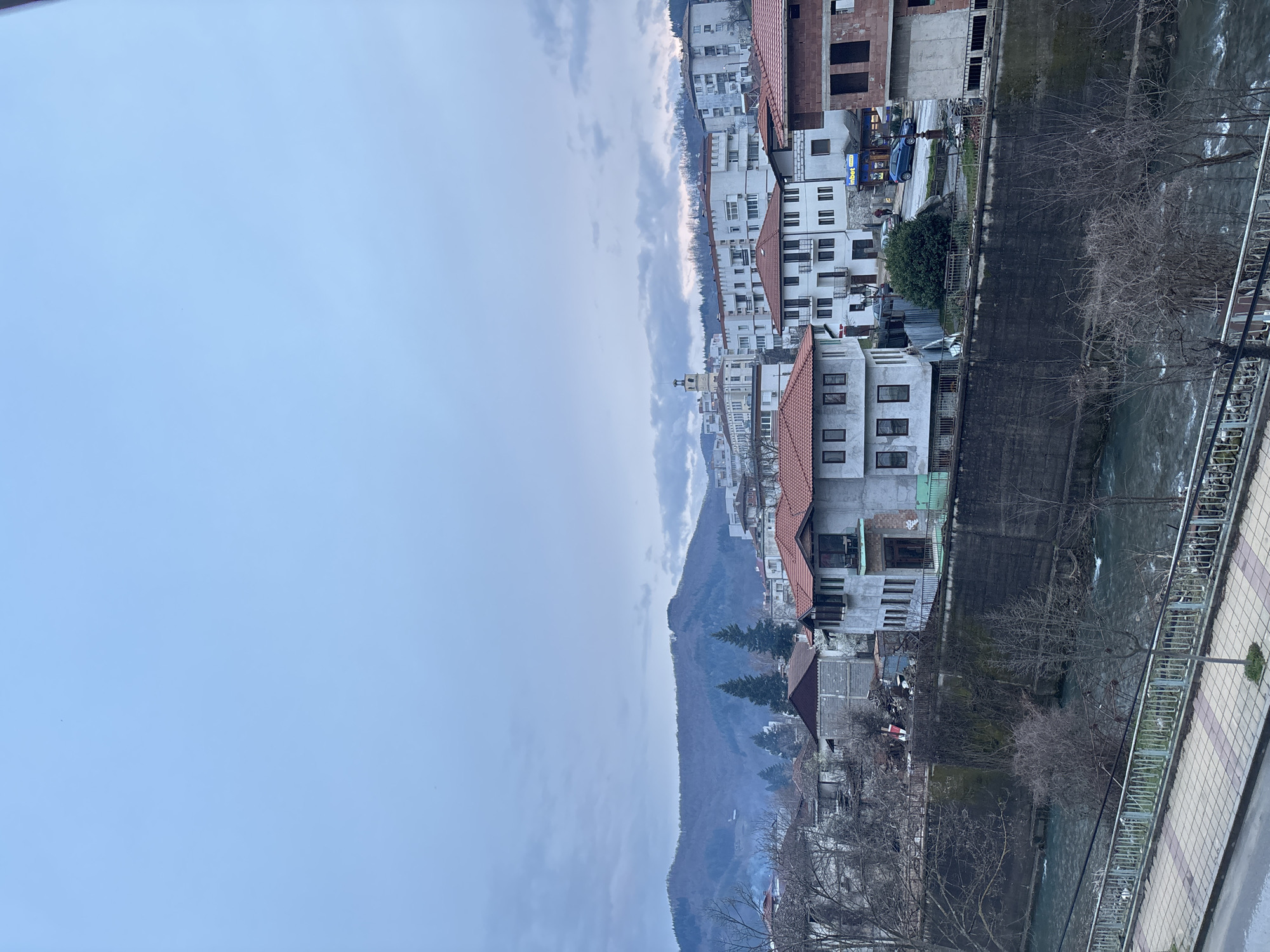 The image is a vertically oriented, slightly tilted view of Zlatograd, Bulgaria, taken from an elevated position, possibly a balcony. The scene is dominated by an overcast sky covering the left half of the image.
To the right, a section of the city stretches into the distance. Rows of buildings, mostly white with red tile roofs, are visible. Some buildings are under construction, revealing brick and concrete structures. A river flows through the city, alongside a pedestrian walkway featuring a metal railing and tiled surface. Bare trees line the riverbank. In the background, a mountain range is partially obscured by clouds.
In the middle, a row of buildings can be seen next to the mountain range. Some of the buildings are under construction, revealing concrete structures.
A blue vehicle can be seen next to the InBet sign.