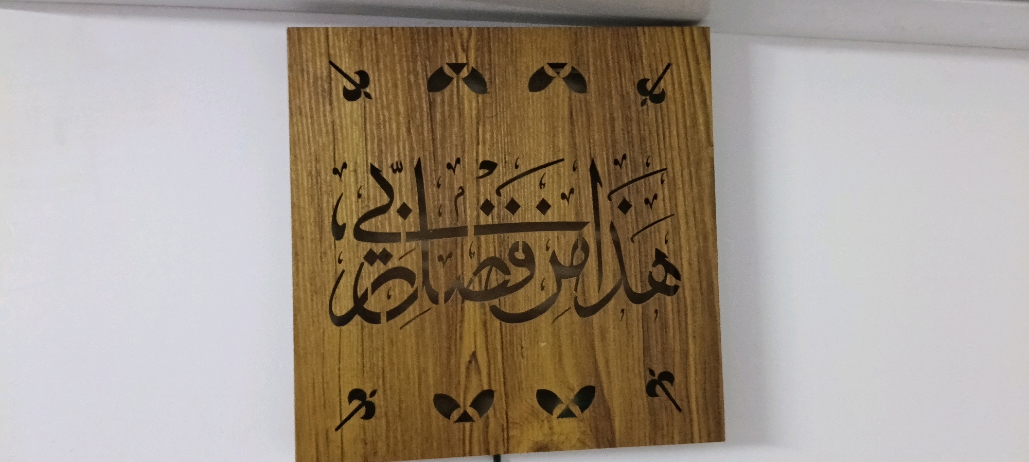 The image shows a rectangular wooden plaque with intricate Arabic calligraphy carved into it. The wood has a visible grain pattern and a warm, light brown color. The calligraphy appears to be black or a very dark color, contrasting sharply with the wood. There are decorative elements in the form of cut-out shapes at the top and bottom of the plaque, resembling stylized leaves or wings. The plaque is mounted on a white wall. The scene is devoid of any identifiable landmarks or features that would indicate a specific location within the city of Udaipur, India. The focus is entirely on the decorative plaque.