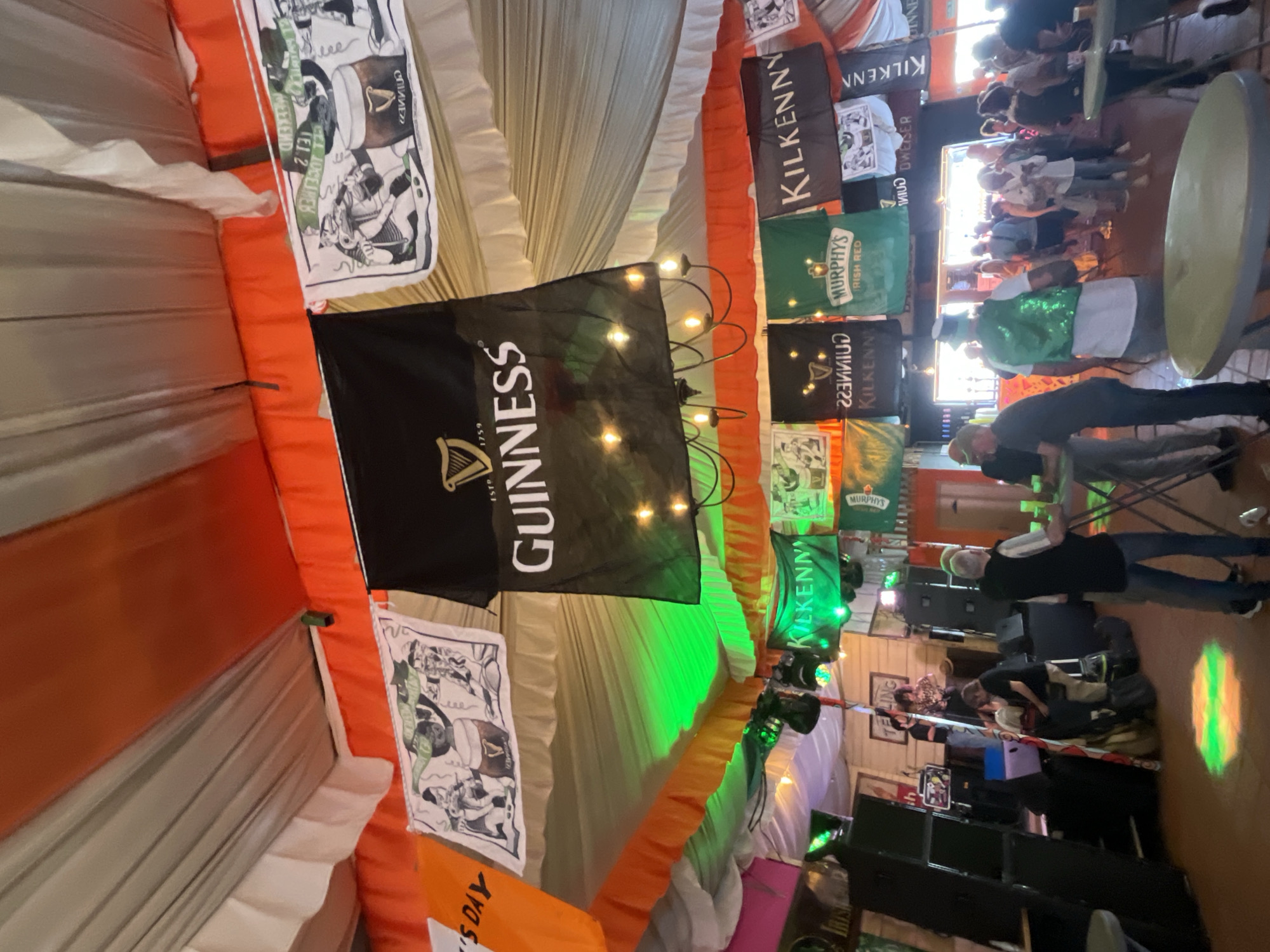 This image captures a lively indoor event, likely a celebration with an Irish theme, set in Fuengirola, Spain. The scene is brightly decorated with orange and white fabric drapes, reminiscent of the Irish flag, adorning the ceiling and walls. Numerous banners for popular Irish brands are prominently displayed, including "GUINNESS," "KILKENNY," and "MURPHYS IRISH RED." Some banners show "1759," associated with Guinness's founding. Illustrated posters featuring people enjoying pints are also visible.
A festive atmosphere pervades the space, illuminated by string lights and green ambient lighting, with a projector casting a decorative pattern on the floor. A group of people are gathered, socializing and standing around tables. One individual stands out in a bright green sequined vest and a matching green top hat, strongly suggesting a St. Patrick's Day celebration. Other attendees are casually dressed, with some appearing to be setting up equipment, including large black speakers and what looks like a sound system, hinting at live entertainment or music. In the background, more people are visible, and light streams in from an opening, indicating it might be daytime outside, despite the indoor lighting creating a cozy, evening-like ambiance. Additional visible text includes "ST" and "SDAY," reinforcing the St. Patrick's Day theme, and "TELLING."