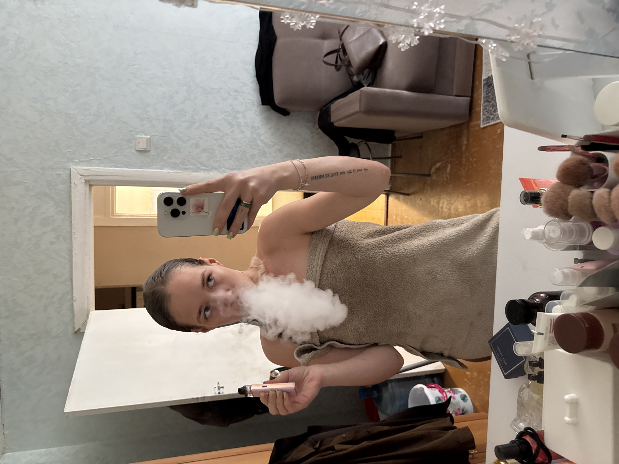 A young woman, possibly a teenager, is taking a selfie in a mirror in what appears to be a bathroom. She is wrapped in a towel and is exhaling a cloud of vapor from a vape pen. Her hair is pulled back, and she is looking at her phone. On the vanity in front of her are various toiletries and makeup items. In the background, a doorway leads to another room, and a chair is visible. The walls are light blue with a textured pattern, and the floor is wooden. The scene is consistent with a residential interior in the city of Chaikovsky, Russia.