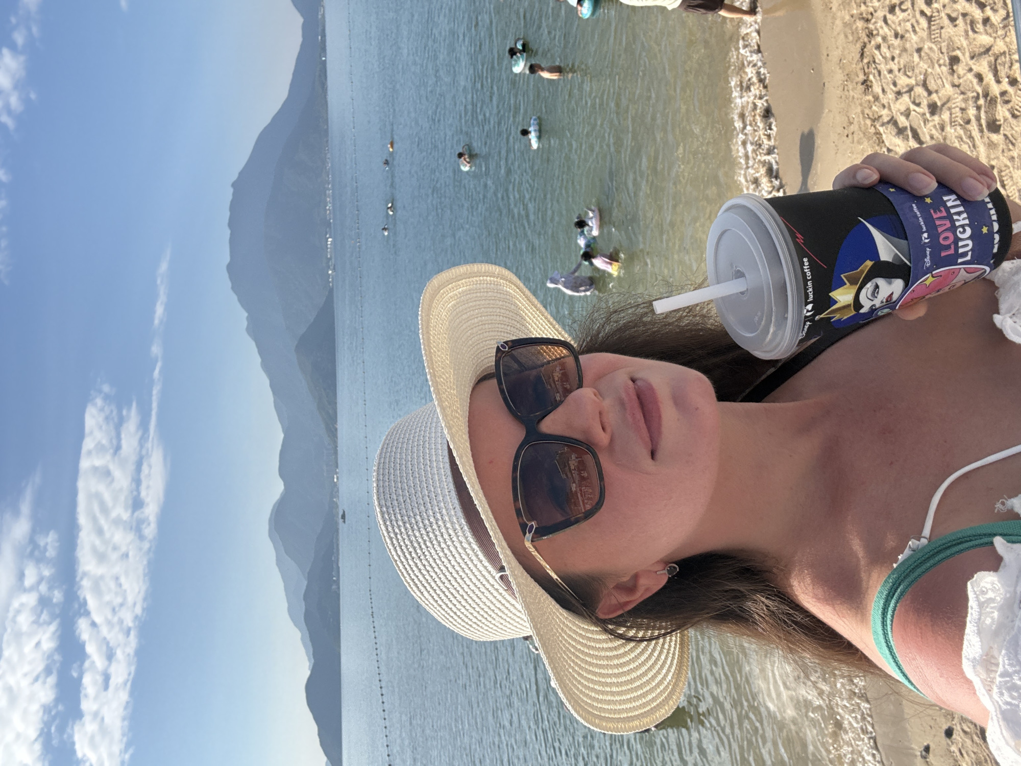 A woman is taking a selfie on a beach in Longgang District, China, holding a Luckin Coffee cup with a Disney villain design. She is wearing a white sun hat, sunglasses, and a bikini top. In the background, the sea is calm with several people swimming and some swimming rings. Mountains are visible in the distance. The sky is clear and blue with scattered clouds. The sand on the beach is light brown.