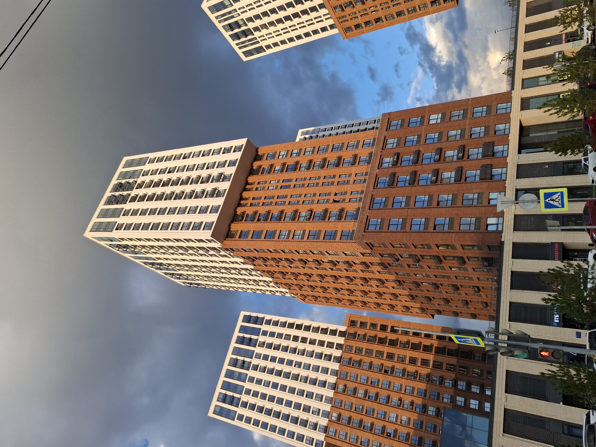 The image depicts a modern residential complex in Tyumen, Russia, under a dramatic sky with dark clouds and patches of blue. Several tall, multi-story buildings dominate the scene. The buildings feature a combination of brick and white facade elements, with numerous windows and balconies. The architectural style is contemporary, with clean lines and a sense of verticality. In the foreground, a traffic light is visible, showing a red pedestrian signal, indicating that pedestrians should not cross. There are also street signs, including a pedestrian crossing sign, and trees lining the street. Several cars are parked along the sidewalk. The overall impression is one of an urban environment with a focus on modern residential architecture. The time of day appears to be late afternoon or early evening, given the lighting conditions and the sky's appearance.
