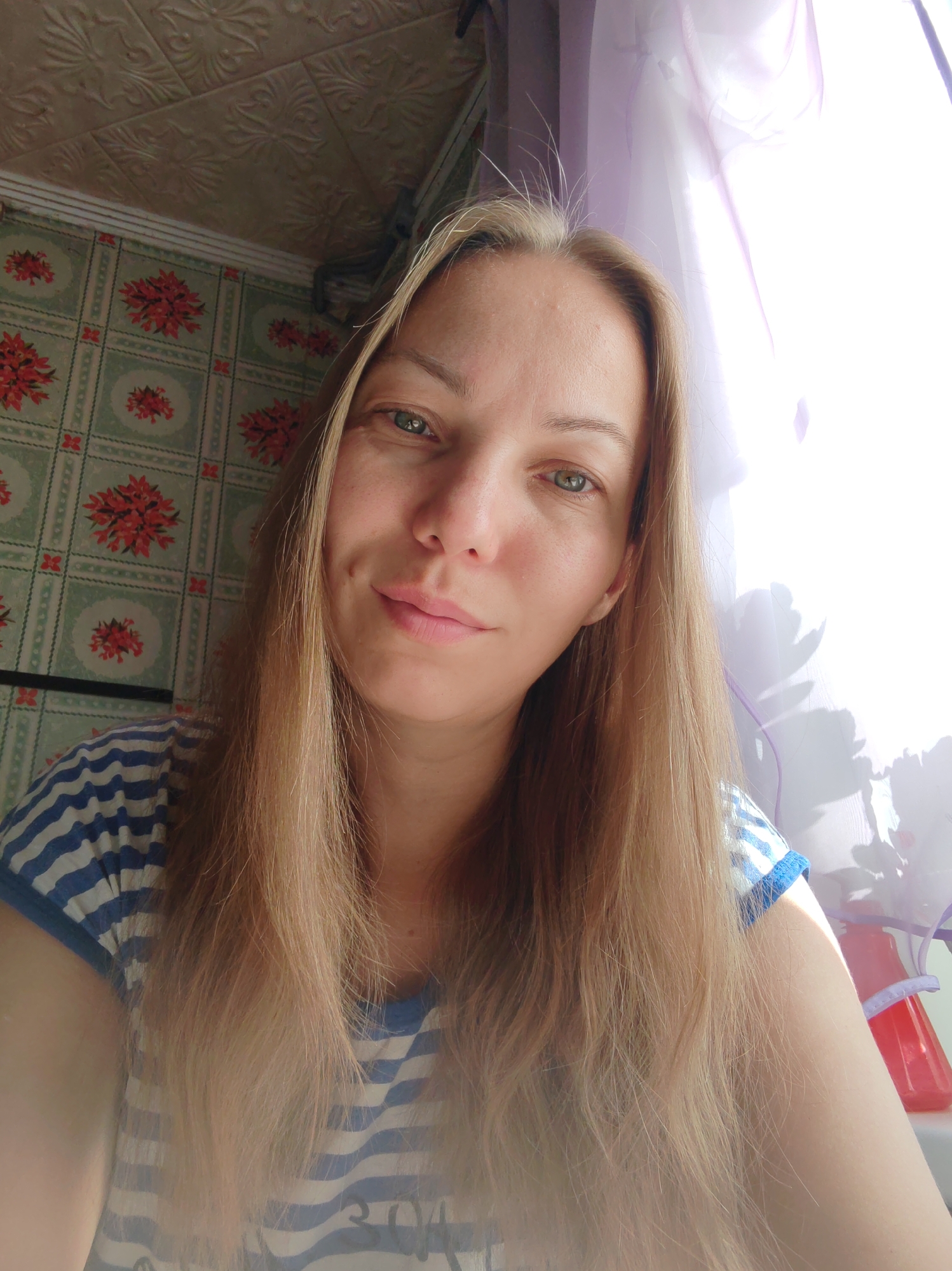 A fair-skinned Caucasian woman with light brown hair and green eyes is pictured in a selfie. She has a dimple on her left cheek and is wearing a blue and white striped t-shirt. The background features floral wallpaper and a bright window with sheer purple curtains, suggesting an indoor setting. The image appears to be taken in the city of Chukhlam, Russia, though no specific landmarks are identifiable.
