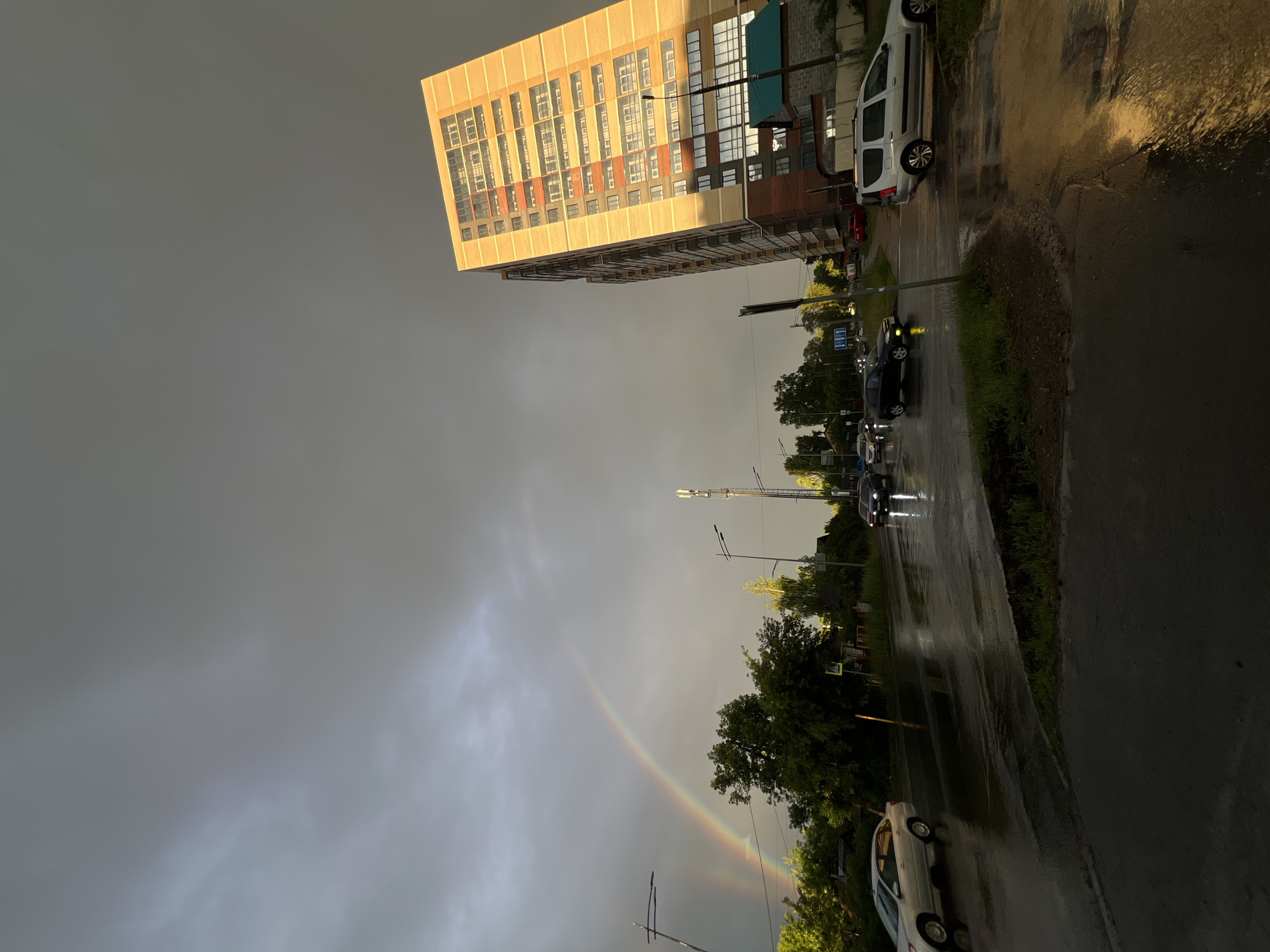 This image depicts a residential street scene in Izhevsk, Russia, under a dramatic sky that shows signs of recent rain and the presence of a rainbow. A tall, modern apartment building with a light yellow facade and a grid of balconies is prominent on the right side of the frame. The street surface is wet, indicating rainfall, and reflections of streetlights and vehicles are visible. Several cars are present, including a silver sedan, a dark-colored sedan, and a silver minivan. A cell tower stands on the right side of the street. Trees and some smaller buildings line the left side of the road, with a faint rainbow arching across the dark, cloudy sky in the distance. The overall atmosphere is moody, transitioning from a storm to clearer skies, with the sun beginning to break through, casting a warm light on the apartment building.
