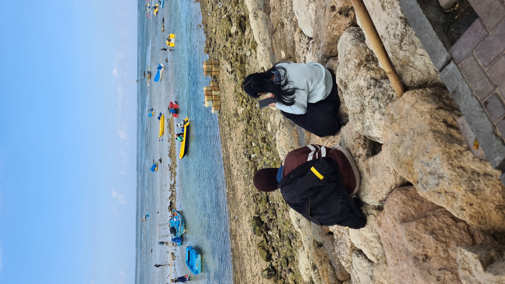 The image depicts a coastal scene in Denpasar, Indonesia, likely near a fishing harbor or a popular beach with boat rentals. In the foreground, two individuals are seated on a rocky embankment, facing away from the camera. The person on the left wears a headscarf and a striped sweater, with a black jacket draped over their shoulders. The person on the right is wearing a light blue sweater and black pants, and is looking at their phone.
The middle ground features a shoreline with scattered rocks and sand. Several small boats, predominantly yellow and blue, are moored or resting on the shallow water and sand. Some of these boats appear to be for recreational use, with people visible in or near them.
The background shows the calm ocean under a clear blue sky. The horizon is marked by a faint line of distant land or structures. The overall atmosphere is one of relaxed activity, with the presence of boats and people suggesting a leisurely or work-related day at the coast. The specific location within Denpasar is likely a coastal area known for its beaches and water activities.