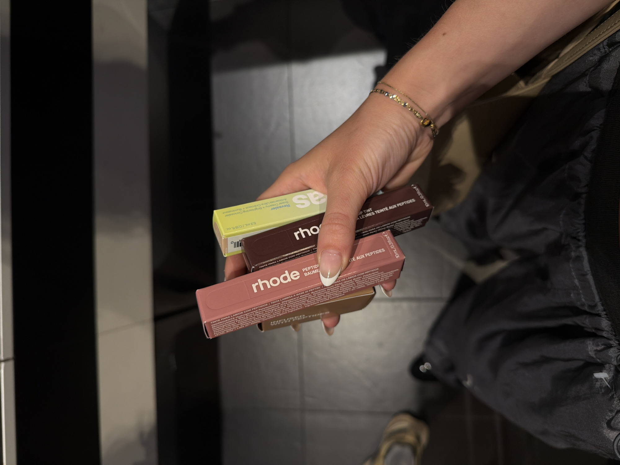 Here is a forensic description of the image:
A person is holding three small boxes of Rhode lip care products. The top box is yellow and labeled "Revealer." The middle box is brown, labeled "LIP TINT, A LEVRES TEINTE AUX PEPTIDES." The bottom box is pink, labeled "rhode PEPTIDE LIP TREATMENT." The person is wearing a gold bracelet. The background includes a black and white striped pattern and gray tile flooring. A black bag is slung over the person's shoulder. The scene appears to be indoors, possibly in a store. Marina del Rey is not explicitly evident in the image.