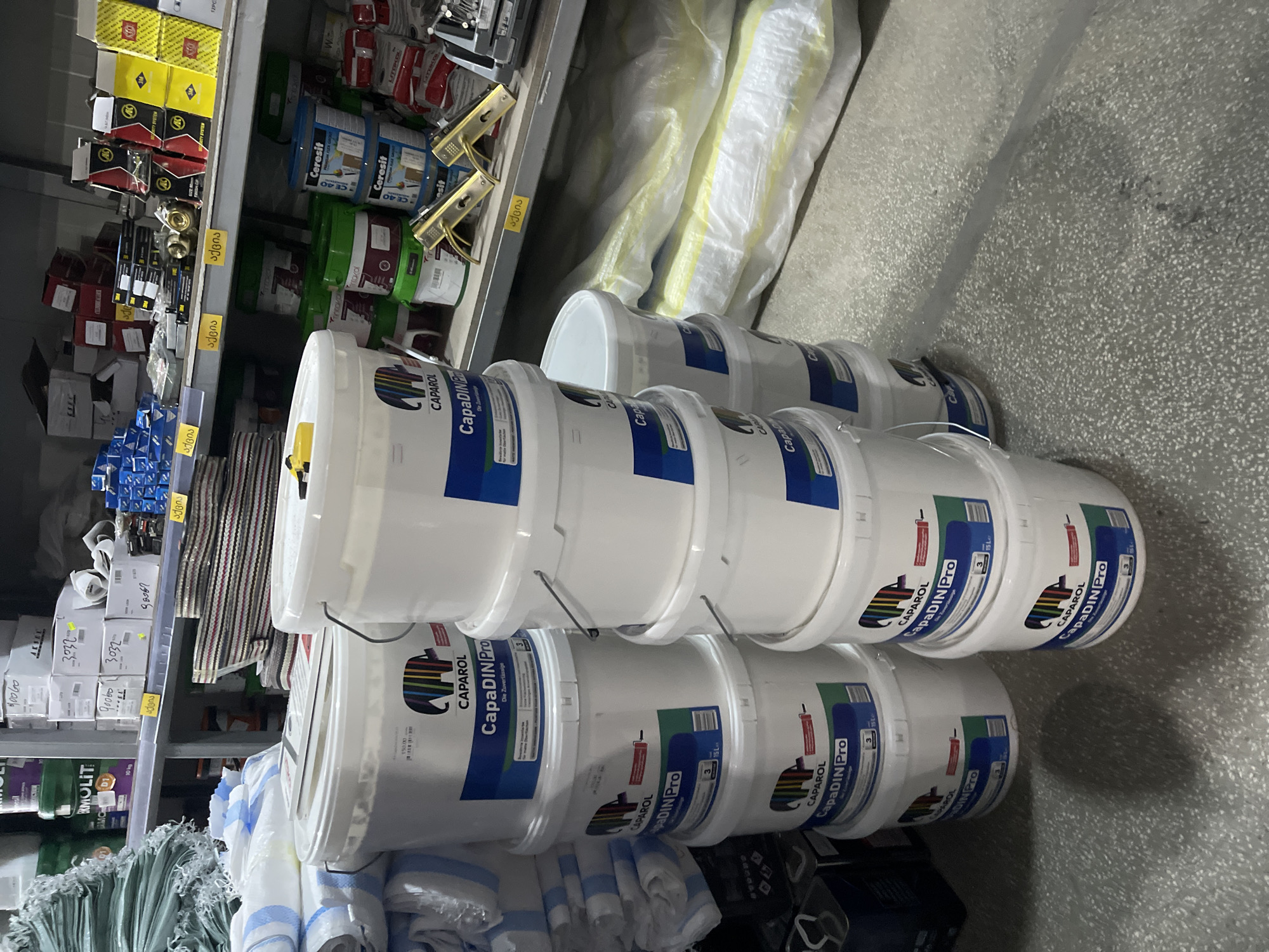 This photograph captures an interior view of a building materials or hardware store, likely situated in Tbilisi, Georgia. In the foreground, two prominent stacks of white "CAPAROL CapaDIN Pro" paint buckets are visible. The left stack contains five 15-liter buckets, while the right stack contains six similar buckets, each featuring a colorful elephant logo. The buckets are marked with "CapaDIN Pro" and "Die Zuverlässige."
Behind these buckets, multiple metal shelves extend across the frame, laden with various products. On the left, several green sacks are stacked. The shelves display an assortment of goods, including green "ERMOLIT" buckets, blue "Ceresit" buckets of various sizes, and numerous white and brown cardboard boxes, some with handwritten labels and yellow sticky notes indicating prices or codes. Smaller packaged items, including hardware components like golden-colored door handles and other metallic parts, are arranged in organized sections. Some product packaging is predominantly blue and red. Yellow price tags or category labels with Georgian text, "აჭივი," are affixed to the shelf edges.
The floor is a light grey concrete speckled with darker aggregate, showing faint linear markings and shadows cast by the stacked buckets. On the lower right, several bags of building material, possibly insulation or mortar, wrapped in white and yellow plastic, rest on the floor beneath a shelf. The store appears well-stocked and brightly lit by overhead artificial lights, creating visible shadows. No individuals are present in the scene.