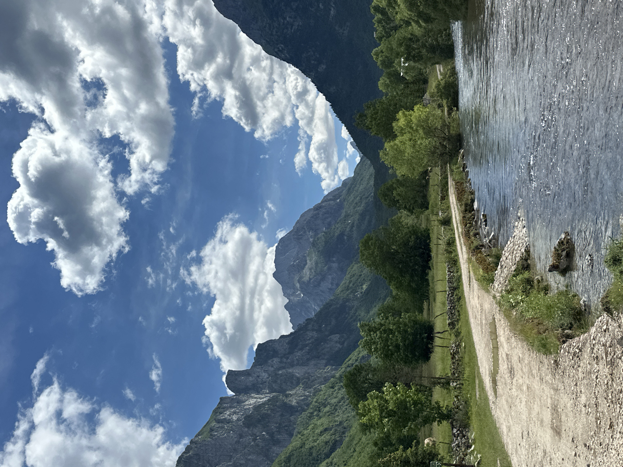 The image depicts a scenic view of a river flowing through a valley surrounded by lush green mountains under a cloudy blue sky. A dirt path runs alongside the river, with a stone wall and some vegetation on the left side. In the background, majestic mountain peaks rise sharply, covered in verdant trees. The river's surface is calm, reflecting the sky and the surrounding landscape. The scene suggests a tranquil natural setting, likely in a rural or mountainous area. Given the location of Gusinje, Montenegro, this image likely captures the natural beauty of the Prokletije mountains, also known as the "Accursed Mountains," which are renowned for their dramatic peaks and pristine valleys. The presence of a river and a path suggests a hiking or natural exploration area. No people are visible in the image.