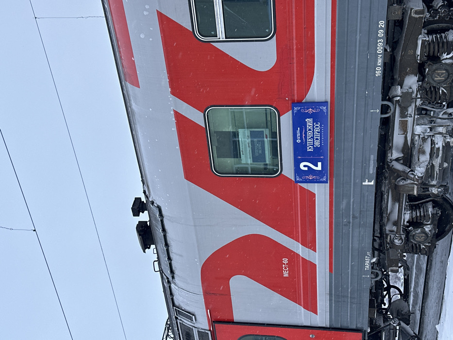 An exterior, mid-shot view captures the side of a grey and red passenger train car, number "2", identified as "КУПЕЧЕСКИЙ ЭКСПРЕСС" (Merchant Express), at an unknown railway location within Unknown, Unknown. Light snow is falling and accumulates on the train's undercarriage and the track below. The train car features prominent red "RZD" branding, multiple rectangular windows, and a blue decorative plaque displaying its name and car number. Additional markings include "MECT-60" (Seat-60), "TAPA57,5T", and "160 KM/Ч 0093 09 20". Snow-covered mechanical components and bogies are visible beneath the train. Overhead electrical lines are visible against an overcast, light grey sky. No individuals are depicted in the scene.
