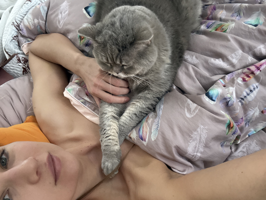 An overhead, close-up image depicts a person and a domestic cat in an indoor setting. The individual, a fair-skinned person with visible light-colored eyes and closed lips, is positioned on the left side of the frame, with their head, exposed left shoulder, and upper arm visible. An orange pillow is partially seen behind their head. Their right hand is gently resting on the torso of a large, short-haired grey cat, which is lying on its left side with its eyes closed, facing the person. The cat's front paws are tucked in. Both subjects are on a bed covered by a light purplish-grey duvet adorned with multi-colored feather patterns. A darker patterned fabric is visible in the upper background. The scene suggests a relaxed interaction in a bedroom environment in Saint Petersburg, Russia.