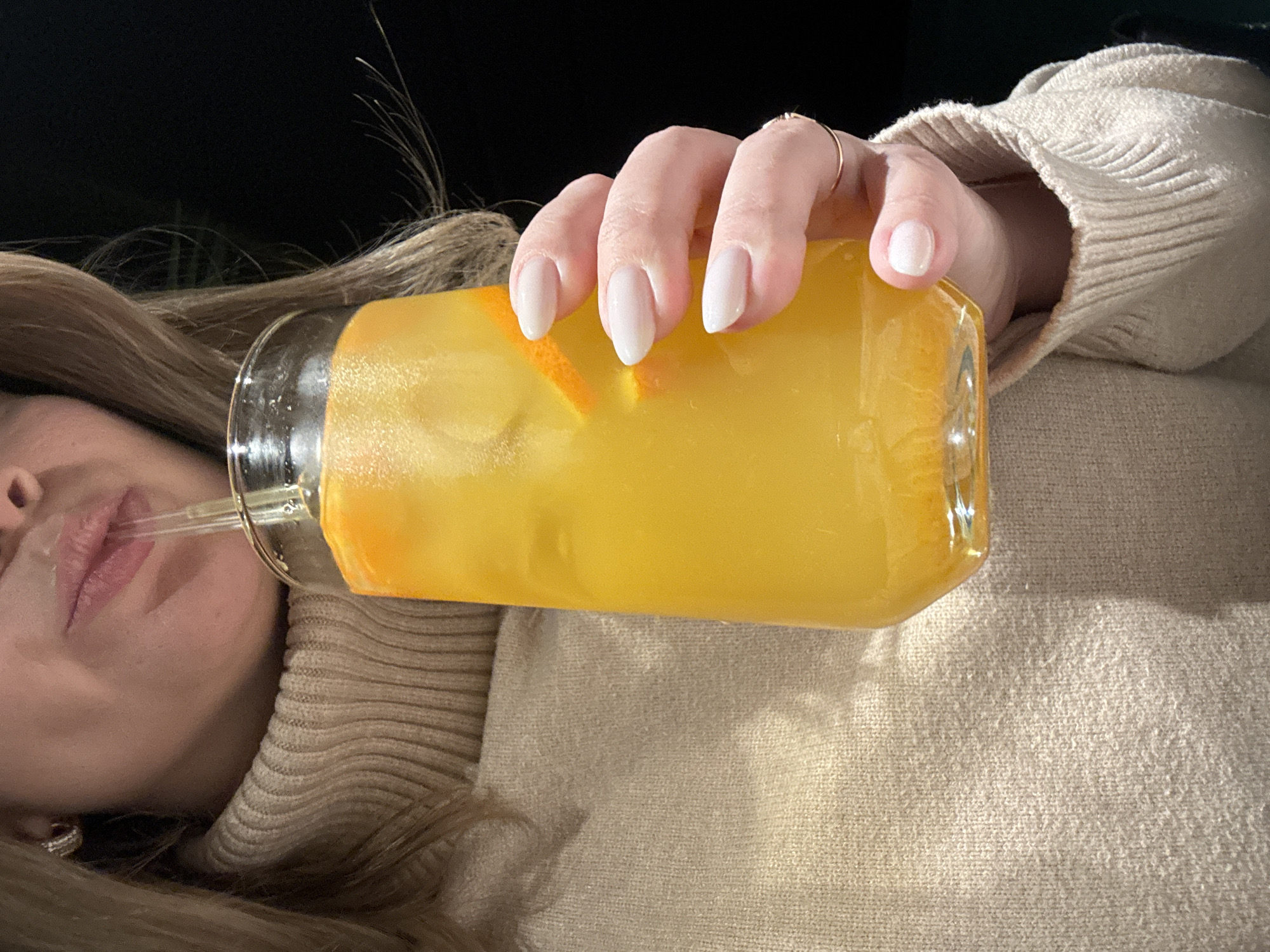 A person is holding a glass filled with a yellow liquid, possibly orange juice or a cocktail, and drinking from it with a straw. The glass contains slices of citrus fruit. The person's left hand is visible, showing white manicured nails and a gold ring on one finger. The person is wearing a beige turtleneck sweater. The background is dark. It is not possible to determine the location within Chaikovsky, Russia, based on this image alone.