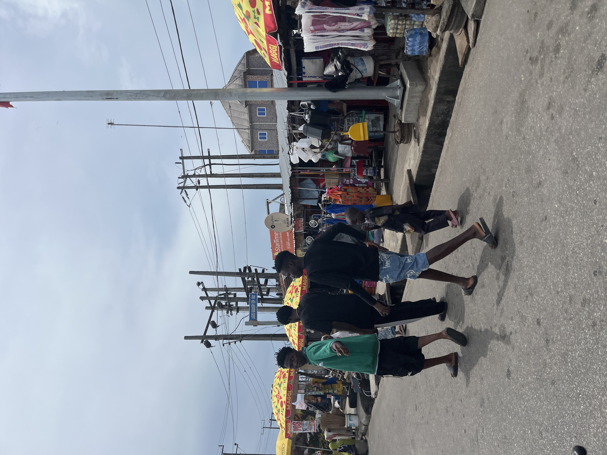 The image depicts three young men walking along a street in Badagry, Nigeria. The street is lined with shops under yellow and red "MAGGI" umbrellas. Overhead are numerous electrical wires and poles. A building with blue-framed windows is visible in the background. Signs for "MTN" and "Starline" are visible, along with street signs for "LAGOS BADAGRY EX".