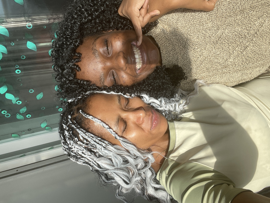 Two women lie side-by-side. One smiles, displaying teeth, and points to her mouth. She has dark, curly hair and wears a tan sweater. The other woman has braided hair that is black and white, and her eyes are closed. She wears a light green top. The background shows a window with a design of leaves and circles. A beam of sunlight illuminates the scene.