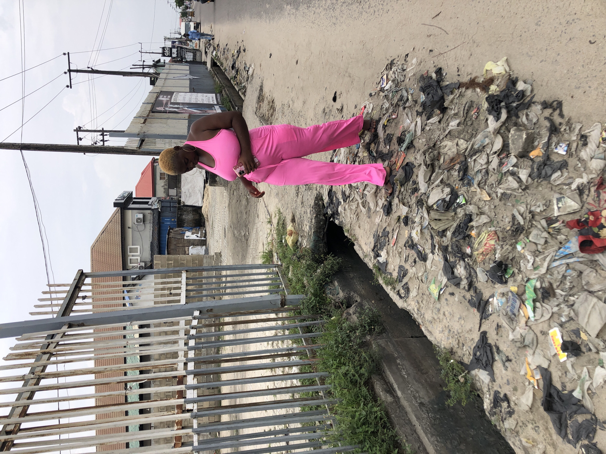 In Ado, Nigeria, a woman in a pink outfit stands on a garbage-strewn sidewalk next to a concrete drainage ditch. She is holding a pink smartphone. The sidewalk is heavily littered with trash of various colors and materials. Behind her is a galvanized metal fence and a building. In the background, utility poles with wires are visible along the street. The sky is overcast.