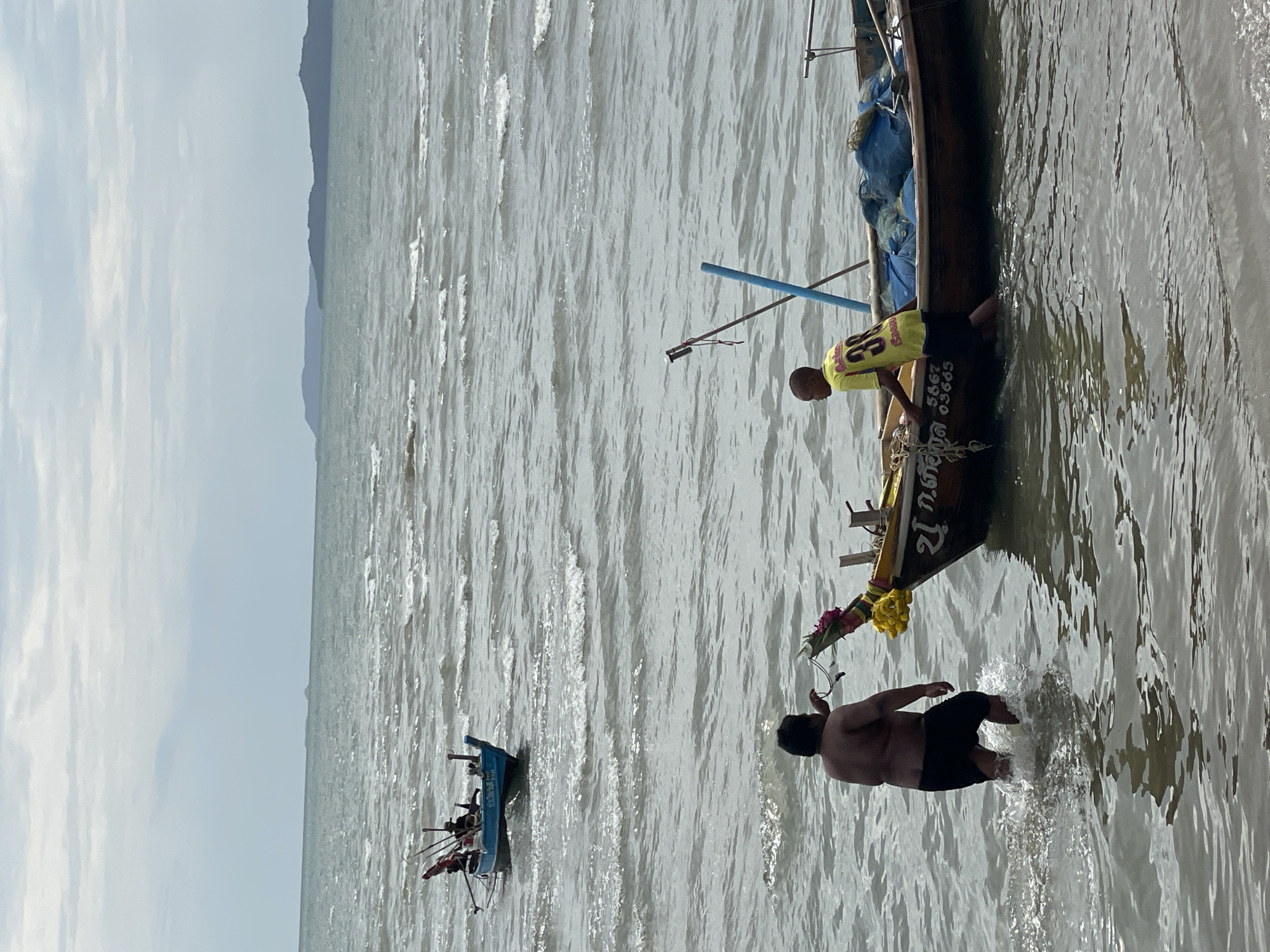 In Ban Na Chom Thian, Thailand, two men are interacting with fishing boats in the shallow ocean. The man on the left, wearing black shorts, is in the water and appears to be holding a rope attached to the boat on the right. The boat on the right, a wooden vessel with a brown hull, has a young man wearing a yellow t-shirt with the number '38' on the back, standing near the front. This boat is decorated with yellow flowers and greenery. There are blue fishing nets visible on board. Further in the background, a smaller blue boat with several people and fishing equipment is anchored in the sea. The water is choppy with visible waves, and distant landmasses are visible on the horizon under a cloudy sky.
