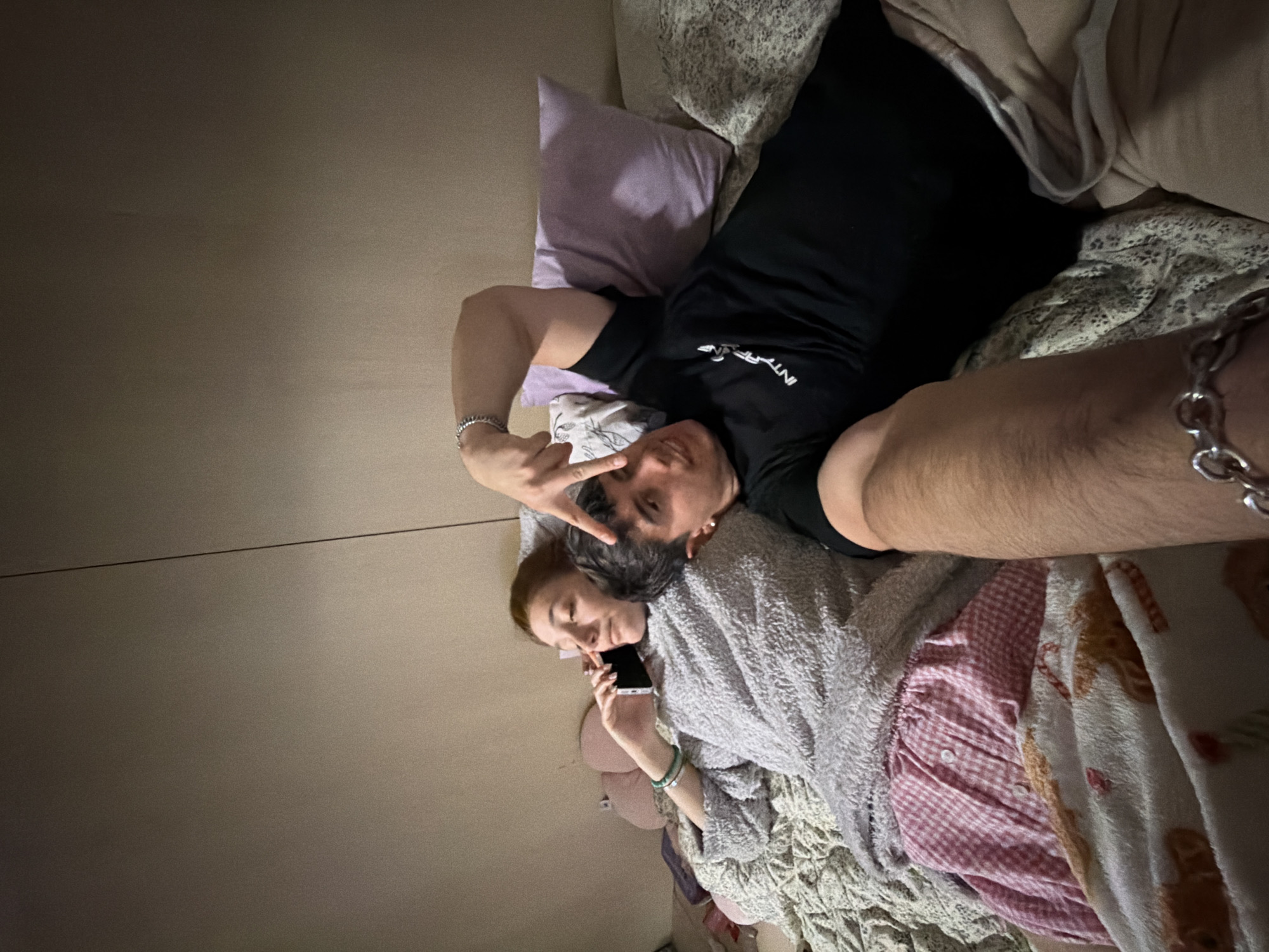 The image captures two individuals, a male and a female, relaxing in a bed indoors, likely in a bedroom in Sofia, Bulgaria. The male, lying on his back, wears a black t-shirt with "INTEЯFONE" or similar white text and a silver chain bracelet on his left wrist. He is making a peace sign with his fingers and looks towards the camera with a slight smile. Beside him, the female is largely covered by a grey textured blanket, with her light brown hair visible. She holds a black smartphone to her ear, appearing to be on a phone call, and wears multiple bracelets on her right wrist, including one with turquoise beads. The bed features a lavender pillow, the grey blanket, a white blanket with light brown/orange patterns, and a pink and white checkered fabric. The background on the left is a plain light brown wall or headboard. The dim lighting suggests the scene takes place in the evening or at night, conveying a relaxed and casual atmosphere.
