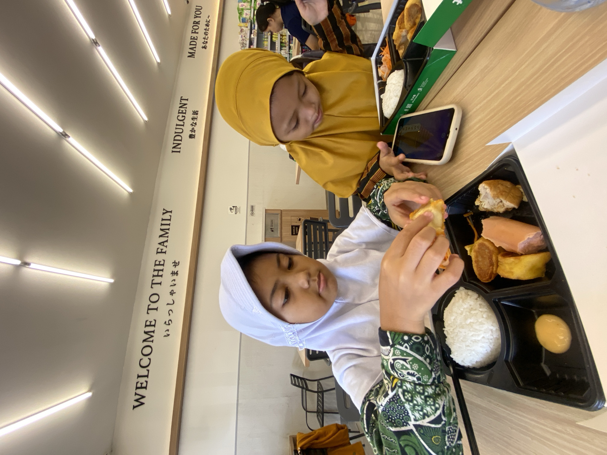 Two young girls are seated at a table in an indoor setting, possibly a restaurant or cafe in Sidoarjo, Indonesia. The girl closest to the viewer wears a white hijab and a green patterned sleeve, holding a piece of food. Her food tray contains rice, a sausage, a fried egg, and other items. The second girl, wearing a yellow hijab, is looking at a smartphone next to her open food container, which contains a similar meal. Text on the wall reads "WELCOME TO THE FAMILY," "INDULGENT," and "MADE FOR YOU" in both English and Japanese. The environment includes interior lighting, tables, chairs, and the presence of another person in the background.