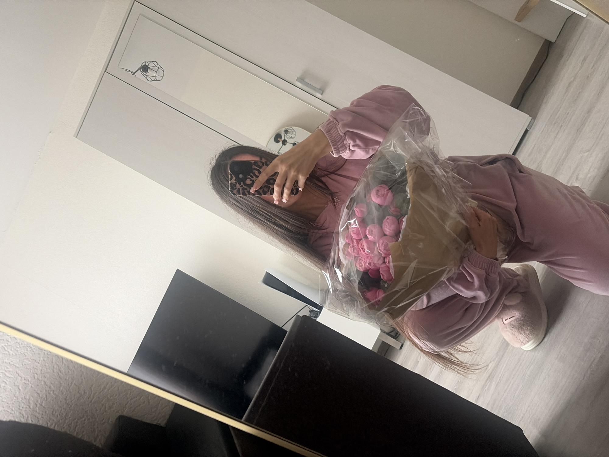 A person wearing pink pajamas and slippers crouches while holding a bouquet of pink flowers wrapped in plastic and brown paper. The person is taking a selfie in a mirror, with their face obscured by a phone case with a leopard print pattern. The background shows a mirrored wardrobe, a dark cabinet, and a television. The floor is light-colored wood laminate.