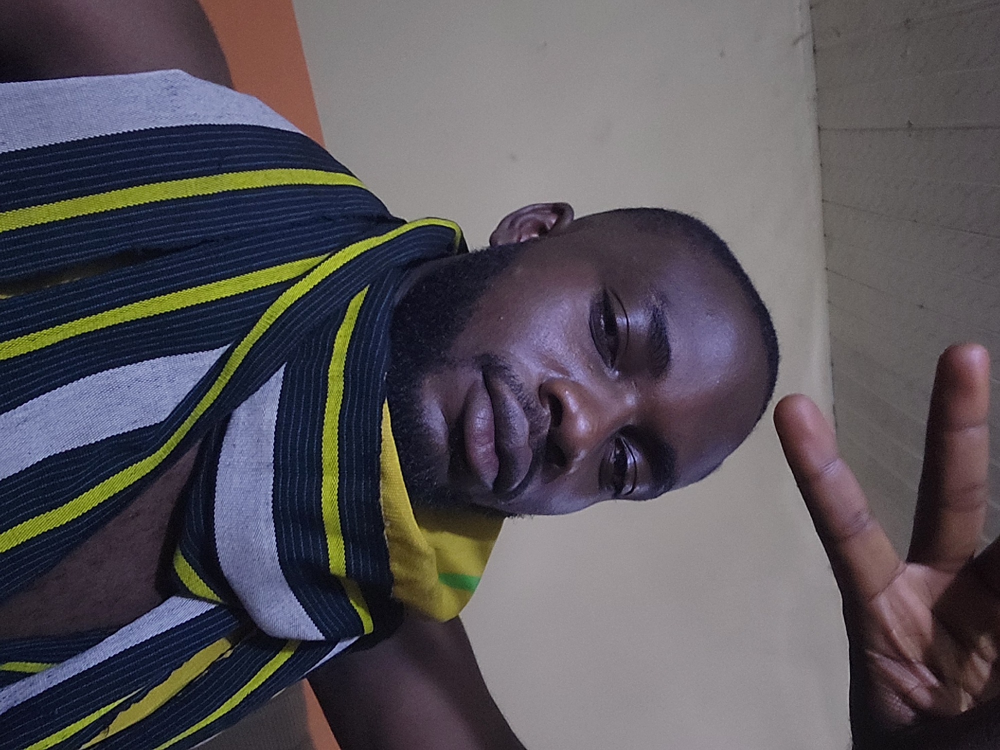 A dark-skinned male subject, estimated to be an adult, is captured in a close-up, head-and-shoulders portrait view, looking directly towards the camera. He has short, dark hair and a neatly trimmed beard. His skin exhibits a slight sheen, particularly on the forehead, nose, and cheeks. The subject is wearing a garment, possibly a scarf or shawl, draped around his neck and shoulders. This garment features a distinctive pattern of horizontal stripes in dark blue/black, bright yellow, and light grey/white. His right arm is raised, with the hand positioned in the upper right quadrant of the frame, forming a "peace" or "victory" sign with the index and middle fingers extended upwards. The background is an indoor setting, featuring a plain, light beige wall in the mid-ground. Above this wall, a light-colored ceiling with visible horizontal seams or paneling is present. The specific location within the city of Jos, Nigeria, cannot be determined from the visual evidence provided in the image.