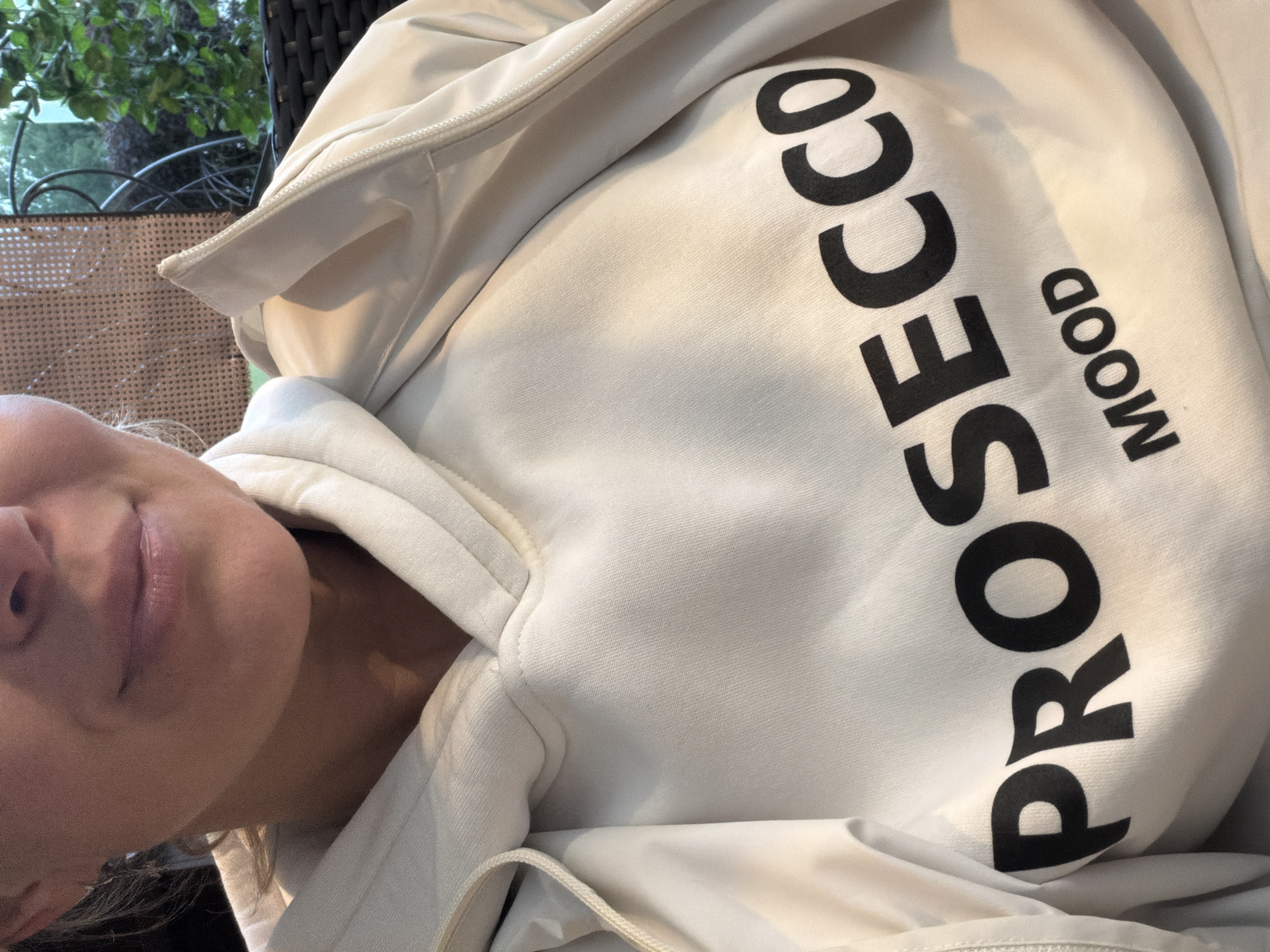 The image presents a close-up, chest-level view of a person seated in an outdoor setting. The person, whose lower face including the chin and lips is visible, has fair skin and appears to be smiling slightly. Light-colored hair is visible near the left ear. They are wearing an off-white or light beige hooded sweatshirt. The front of the sweatshirt prominently displays the word "PROSECCO" in large, arched black capital letters, with "MOOD" in smaller, straight black capital letters centered underneath. Over the sweatshirt, a light-colored (possibly white or very light grey) jacket or coat is worn unzipped, revealing a zipper detail along its opening on the right side.
In the background to the left, a portion of a light brown, woven material, possibly rattan or wicker from a chair back or screen, is visible. To the right, green foliage and a dark, curved metal structure (likely part of outdoor furniture or a railing) indicate an outdoor environment such as a patio or garden. No visual cues within the image identify its specific geographic location, including the city of Chaikovsky, Russia.