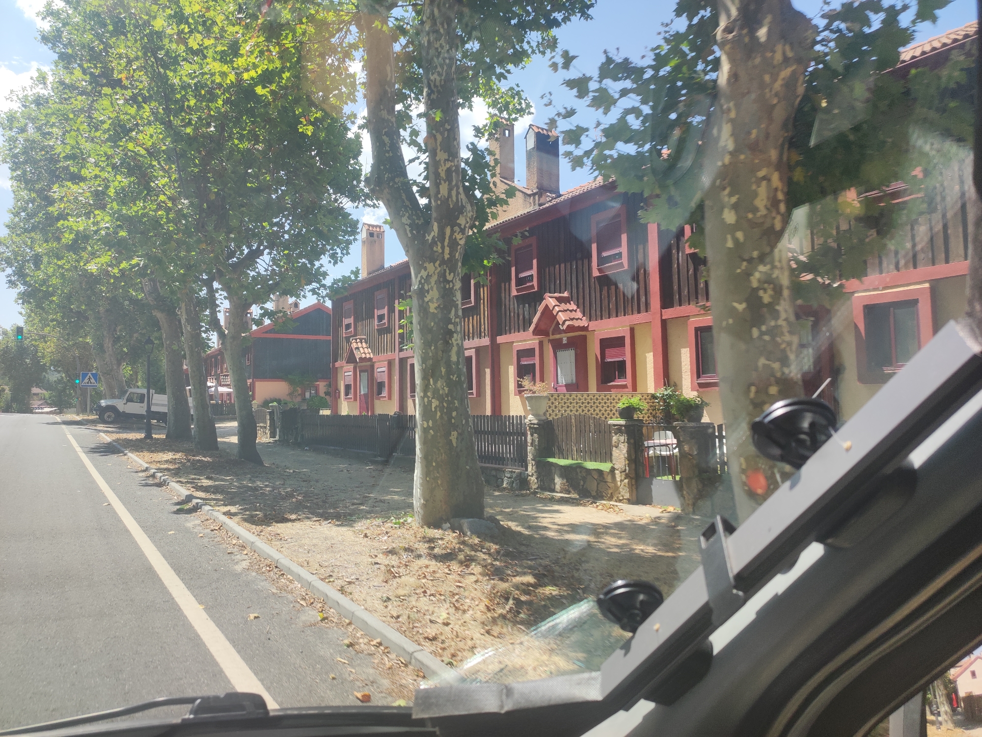 The image shows a street scene in Real Sitio de San Ildefonso, Spain, on a sunny day. The viewpoint is from inside a vehicle, looking out through the windshield. A paved road runs diagonally from the bottom left towards the middle of the frame, marked with a white line. To the left of the road is a sidewalk with a curb. A white pickup truck is parked on the right side of the road, near a pedestrian crossing sign.
A row of large trees with green foliage lines the right side of the road, casting shadows on the ground. Behind the trees, a series of attached houses with red tile roofs and wooden facades painted in shades of brown and yellow are visible. These houses have multiple windows with red shutters and chimneys. A wooden fence runs along the front of the properties. The ground is covered with dry leaves and scattered debris.
In the foreground, the interior of the vehicle is partially visible, including parts of the dashboard, windshield wipers, and mounting brackets with suction cups, likely for a device. The overall impression is a quiet residential street in a town during daylight hours.