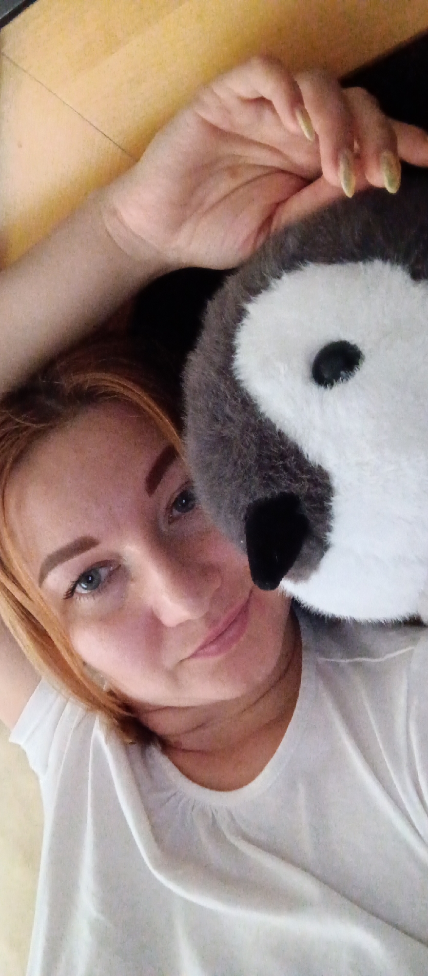 The image shows a woman with fair skin and light red hair lying down. She is wearing a white T-shirt and has blue eyes. To her right is a stuffed penguin toy. The background appears to be a light brown wooden surface, possibly a floor. The location appears to be indoors.