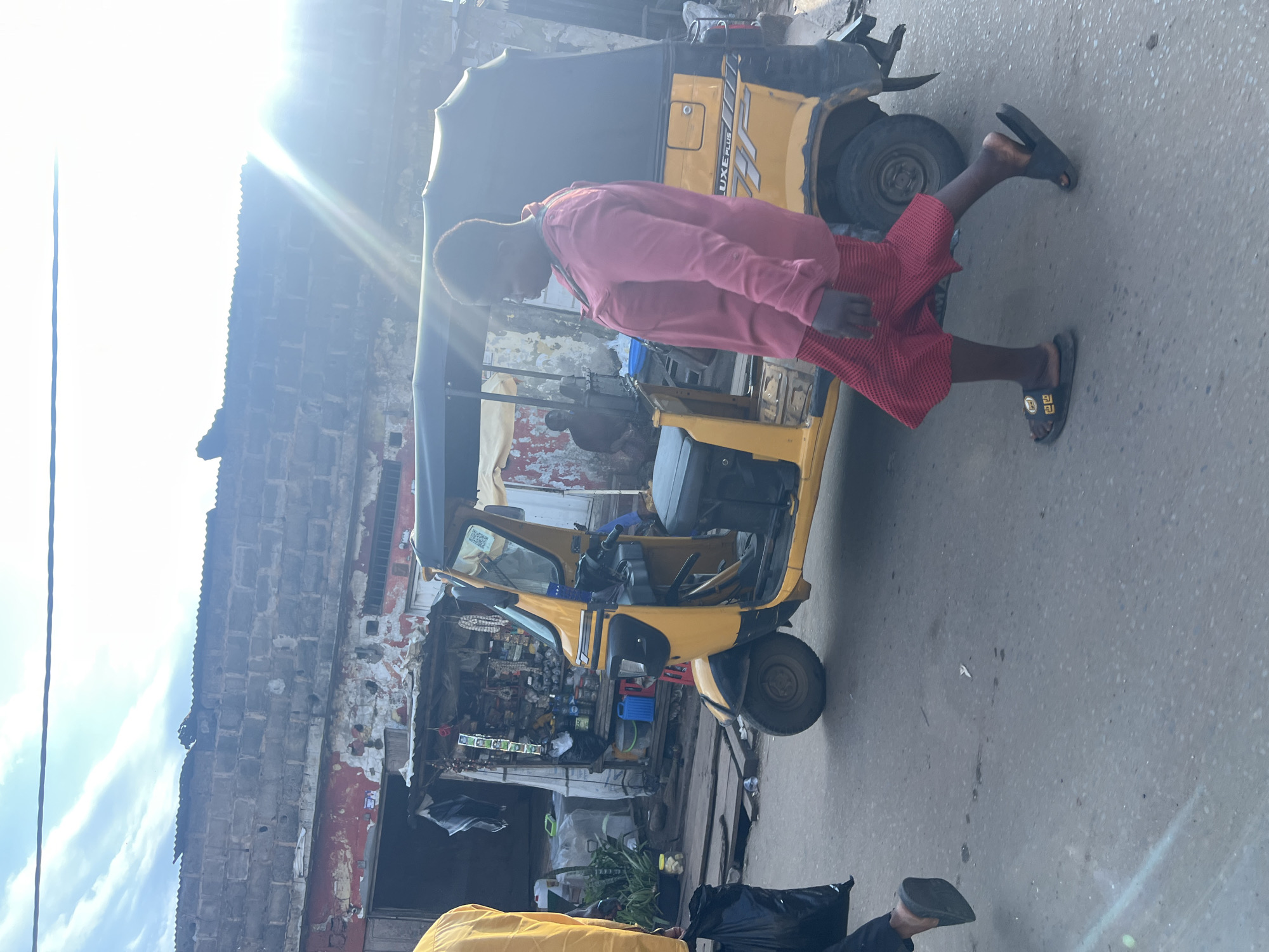 This media file shows a scene in Nkpor, Nigeria, likely during the daytime, indicated by the bright sunlight causing lens flare. The primary subjects are a person and a yellow auto-rickshaw, also known as a "keke napep." The person, wearing a pink long-sleeved shirt and red dotted shorts, is walking on the right side of the road, with their body angled towards the auto-rickshaw. They are wearing black slides with gold accents.
The auto-rickshaw is parked on the left side of the frame, appearing to be a mode of transport or a business stall, as there are items displayed within its open structure, suggesting a roadside shop or market stall. The background features a brick building with peeling paint and a corrugated roof, characteristic of many urban areas in Nigeria. There's also a glimpse of a blue bucket and black trash bags.
The overall scene depicts a typical street environment in Nkpor, with elements of commerce and daily life. The sunlight suggests it is likely midday or early afternoon. The image is oriented vertically, and the bright light from the sun is a prominent feature, creating a strong contrast and some overexposure.
