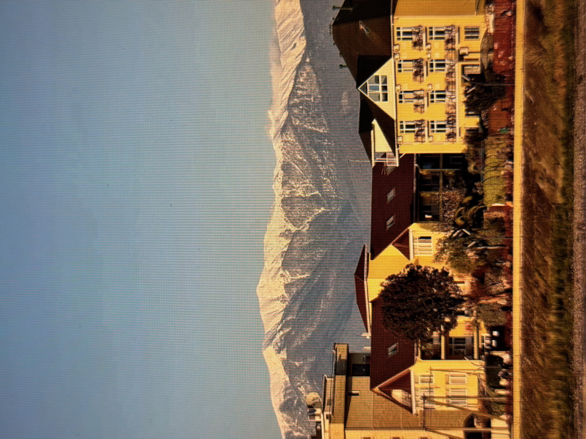 The image depicts a scenic view with a clear blue sky on the left, transitioning to a majestic snow-capped mountain range in the middle. The mountain's rocky slopes are textured and illuminated by what appears to be late afternoon or early morning sun, casting long shadows and highlighting the rugged terrain. On the right side of the image, several buildings with yellow and brown facades are visible, featuring numerous windows. These buildings are nestled in front of the mountains, suggesting a town or village situated at the foothills. Lush green trees are scattered between the buildings, adding a touch of nature to the urban landscape. The overall setting evokes a sense of peaceful coexistence between human habitation and the grandeur of nature. The image appears to be taken from a slightly elevated perspective, possibly from another building or a nearby hill. The lighting suggests a time of day when the sun is low in the sky, creating warm tones on the mountains and buildings. There is no visible text in the image.
