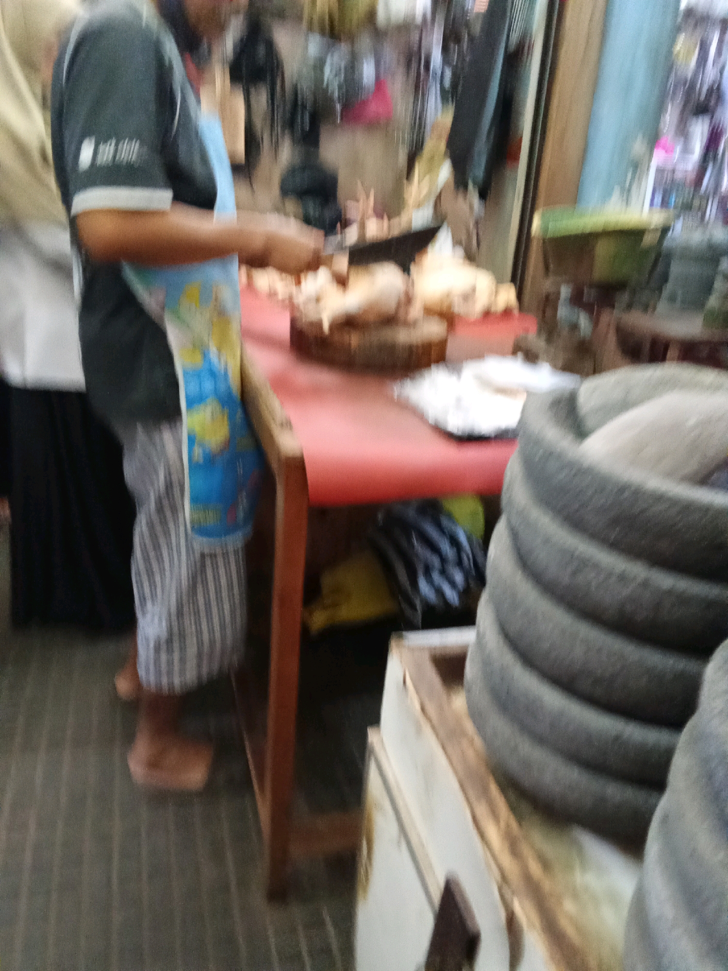 The image depicts a person in a market stall, likely in Banjarnegara, Indonesia, preparing food. The person, wearing a dark shirt and a patterned apron, is positioned behind a red-covered counter. Their hands are visible, suggesting they are engaged in a task involving food preparation, possibly cutting or arranging. On the counter, there are several whole chickens and what appear to be wooden cutting boards, indicating a butcher or food vendor setting. In the foreground, to the right, a large, stacked stone mortar or grinder is prominently featured, a common item in Indonesian kitchens and markets. The background is blurred but shows market stalls with various goods, including bags and hanging items, typical of a local Indonesian market environment. The lighting suggests it is daytime, possibly indoors or under cover within the market. The overall scene conveys a sense of a busy, functional marketplace.