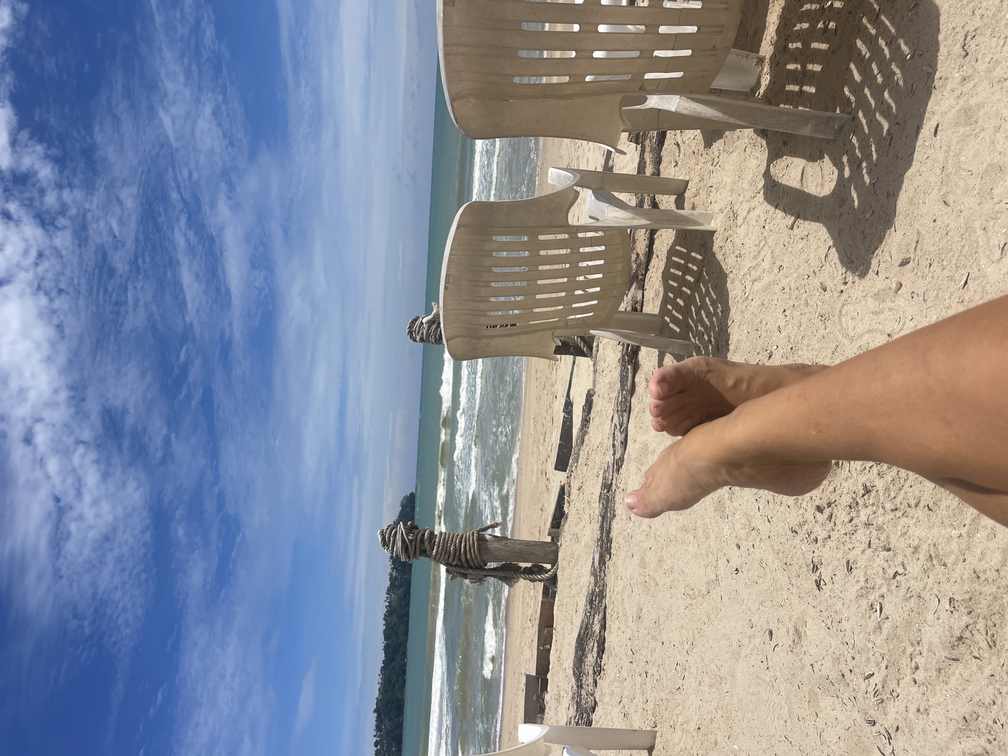 This image depicts a beach scene on a sunny day. In the foreground, a person's legs and feet are crossed, resting on the sand. Behind them, two white plastic beach chairs cast shadows on the sandy ground. Further back, the ocean stretches out to the horizon, with gentle waves rolling onto the shore. A wooden post wrapped with rope stands near the water's edge. In the distance, a green, tree-covered hillside with buildings is visible. The sky is a vibrant blue, dotted with scattered white clouds. The overall environment suggests a tranquil beach setting in Choeng Thale, Thailand.