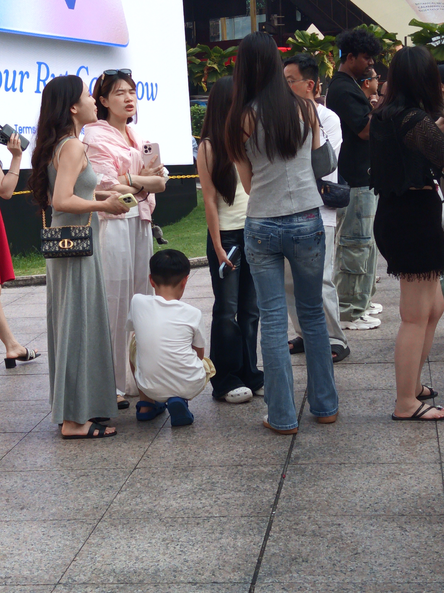 The image captures a group of people standing on a paved outdoor area, possibly a pedestrian walkway or plaza in Kuala Lumpur, Malaysia. In the foreground, a young boy in a white t-shirt and blue shorts is squatting down. To his left, a woman in a long grey dress with a designer handbag is standing. Next to her, a woman in a pink top and white pants is holding a smartphone and looking up and to the left, seemingly engaged in a conversation or reacting to something. Other individuals are visible in the background, including a man with curly hair and a black shirt. The setting appears to be urban, with a large screen or billboard visible in the upper left portion of the image. The ground is paved with textured tiles.
