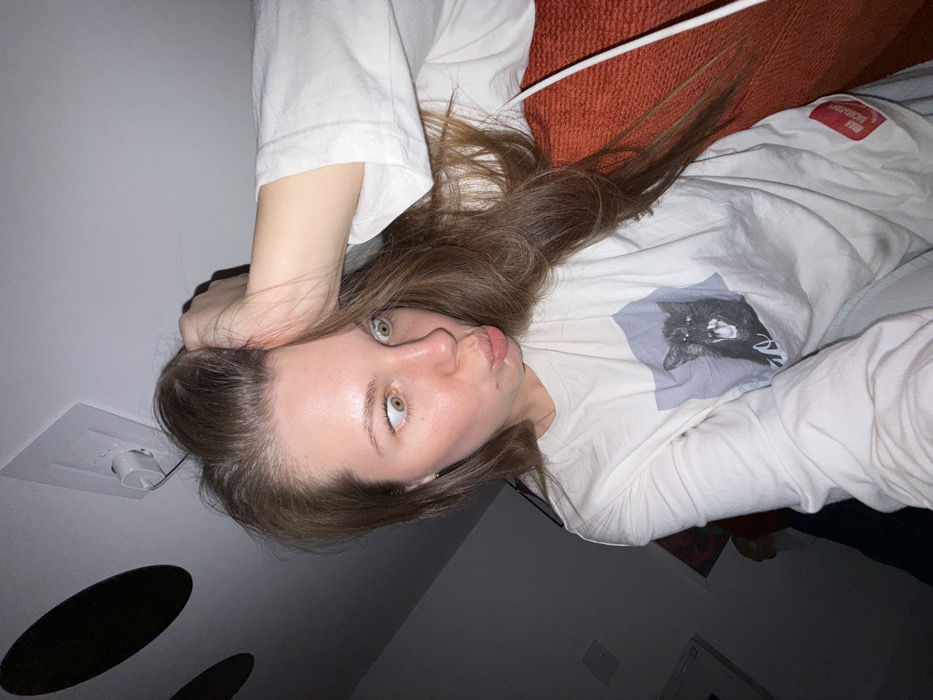 The image displays a young adult female indoors, captured in a self-portrait. She possesses long, light brown hair, with her right hand positioned on top of her head, fingers splayed in her hair. Her eyes are wide and appear slightly convergent, exhibiting green irises. Her lips are puckered into a distinct "fish-face" expression. She is attired in a long-sleeved, off-white t-shirt featuring a prominent graphic of a black cat with an open mouth, displaying teeth, against a light grey rectangular background. A red rectangular label with partially visible white text, including "HMA LABORATORI," is present on the lower portion of her left sleeve. The background is dimly lit, revealing an interior space. A white electrical outlet with a white charger plug inserted is visible on a white ceiling or wall in the upper left. Two dark, circular objects are partially discernible on the ceiling/wall. To the right of the subject, a textured, orange or rust-colored fabric surface, consistent with upholstery, is present, with a white cable running vertically against it. The specific location within the city of Vozdovac Urban Municipality, Serbia, is not discernible from the visual content of this image.