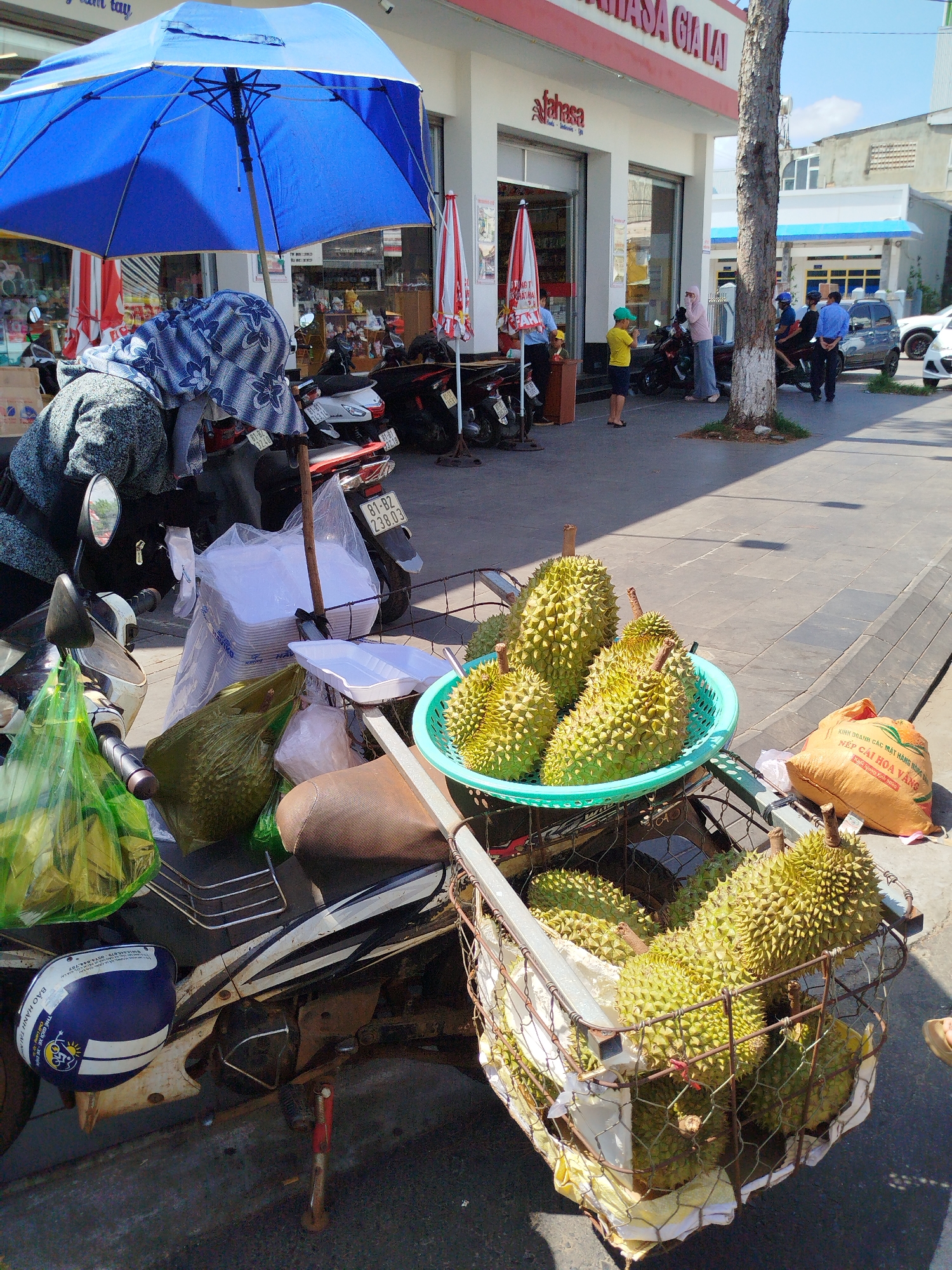 This image shows a street scene with a parked scooter displaying durian fruit for sale in Pleiku Ward, Vietnam. The scooter, white with black accents, is heavily loaded with a blue helmet attached to the side, bags of produce, stacks of white food containers, and a wire basket overflowing with durian. A turquoise plastic bowl also contains durian. A blue umbrella is positioned above and behind the scooter, presumably to provide shade. Behind the scooter, there are other parked scooters and the facade of a building labeled "Lahasa Gia Lai." In the background, there are people, including a child wearing a green hat and a small group of people talking. A car is also visible. The street appears paved and the overall lighting suggests it is daytime. The image focuses primarily on the durian fruit and the scooter.
