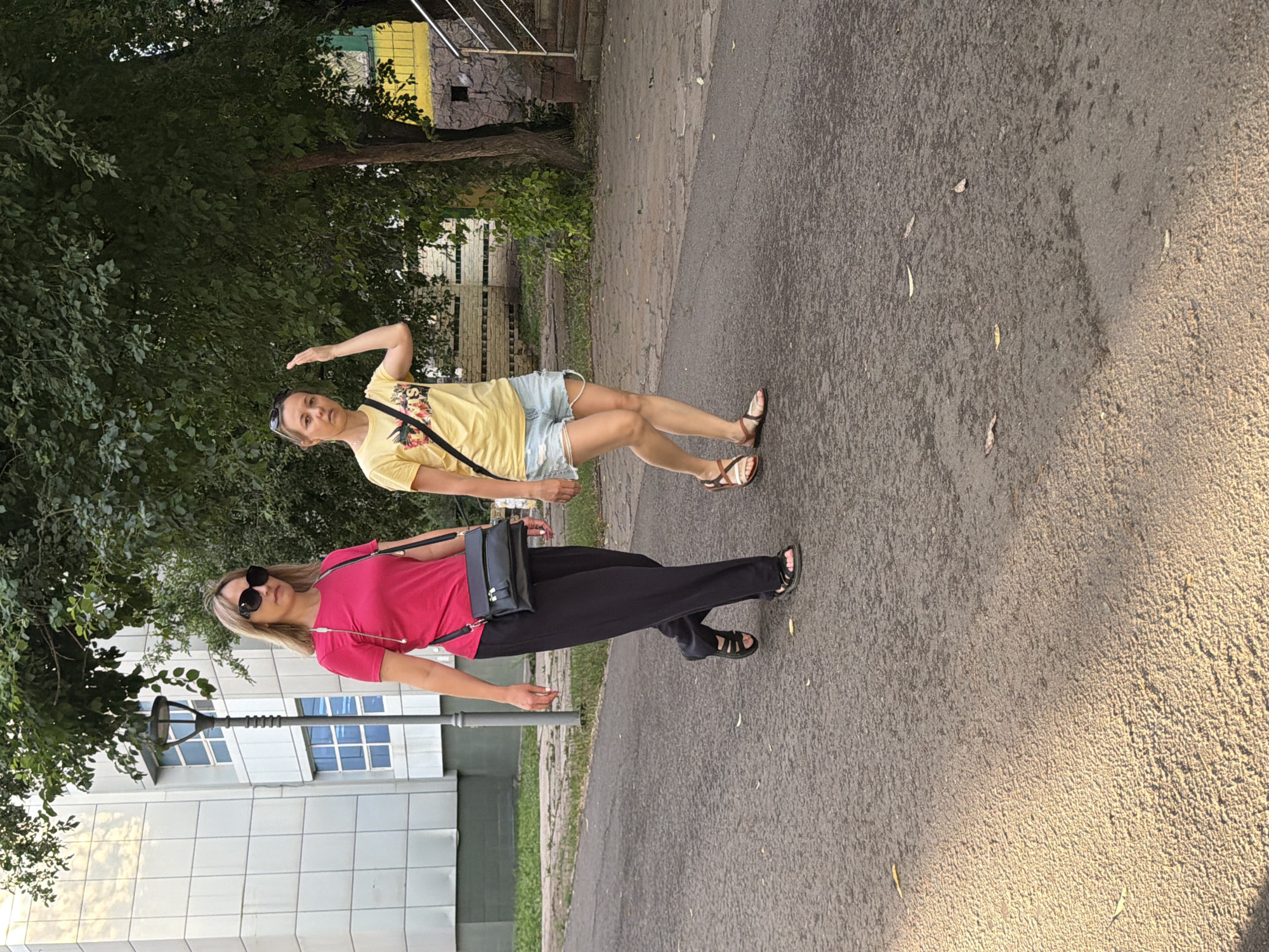 Two women are walking on a paved street in Astana, Kazakhstan, near a building. The woman on the left is wearing a bright pink t-shirt, black pants, black sandals, and sunglasses. She has a black crossbody bag. The woman on the right is wearing a yellow t-shirt, denim shorts, and brown sandals. She is shielding her eyes from the sun with her hand. The background includes a modern building with white tiled facade and blue-trimmed windows, a street lamp, and a leafy tree. The street surface appears damp in some areas, possibly from recent rain or watering.