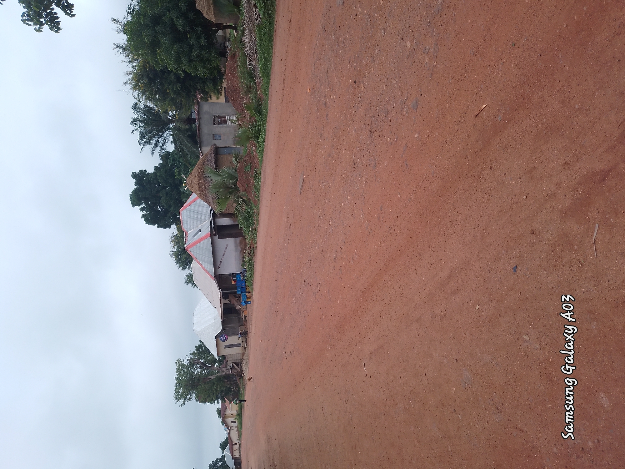 A wide, dusty, reddish-brown dirt road stretches across the foreground of the image. In the background, several structures line the side of the road. These include a few small, simple houses with corrugated metal roofs, some with thatched roofs. A small, light brown dog stands near the middle of the road, facing left. A person in a green shirt and dark pants walks away from the camera on the left side of the road. A few palm trees and other green foliage are visible behind the buildings, suggesting a rural or semi-rural setting in Doka-Tofa, Nigeria. The sky is overcast and a pale gray. The bottom left corner of the image contains a watermark that reads "Samsung Galaxy A03".