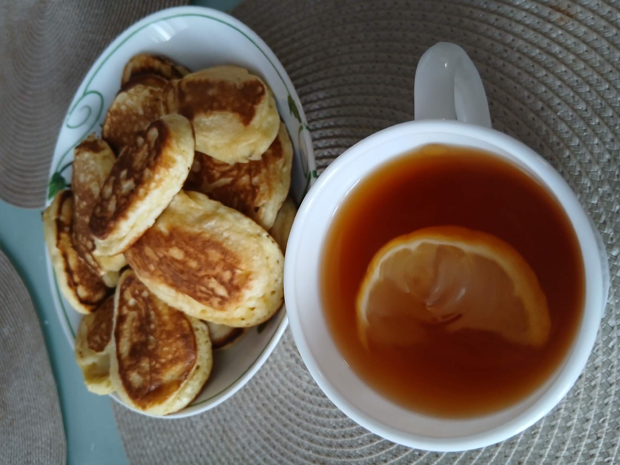 This image shows a close-up view of a breakfast or snack. The primary objects are a bowl of small, golden-brown pancakes and a white mug filled with amber-colored liquid, likely tea, with a slice of lemon floating on top. The pancakes are stacked in the bowl, which has a white rim with a green decorative pattern. The mug is partially visible on the right side of the frame, and its handle is clearly seen. Both the bowl and the mug are placed on a woven placemat with a circular pattern. The background is a light blue surface, possibly a table.
The scene depicts a simple, domestic setting, likely a kitchen or dining area. The lighting appears to be natural, suggesting it could be daytime. There are no people visible in the image. The overall mood is relaxed and inviting, suggesting a moment of quiet enjoyment. There is no visible text in the image. The mention of "Chaikovsky, Russia" provides a geographical context for the scene, implying it was taken in that location.