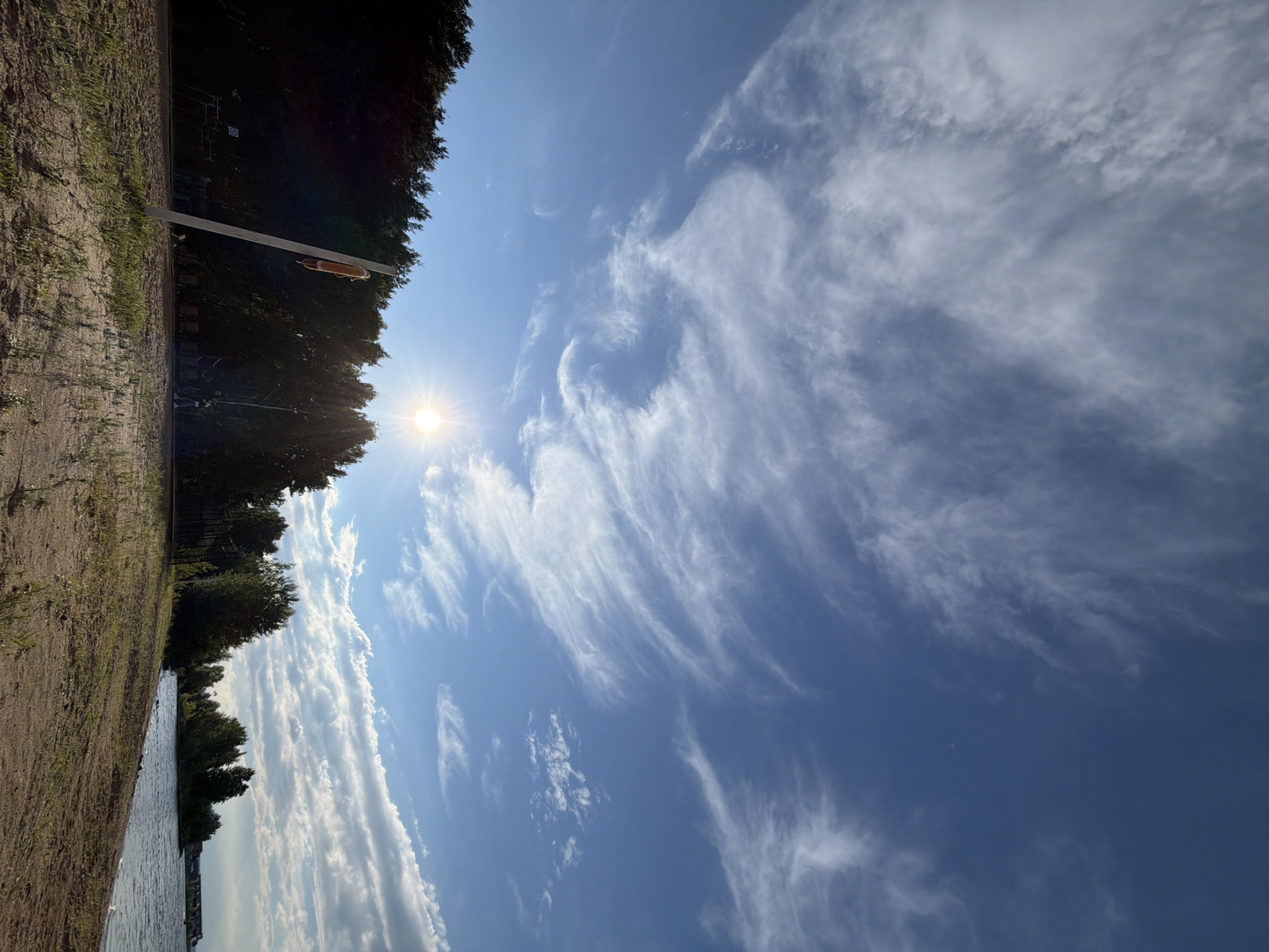 The image shows a wide-angle view of a sandy beach on a sunny day. The sky is a vibrant blue, with scattered wispy white clouds. The sun, a bright orb with visible rays, is positioned high in the sky, casting a warm glow over the scene. In the foreground, a sandy path leads towards a line of dark green trees that form a dense forest. A white pole with an orange life preserver attached stands prominently on the left side of the beach, angled towards the sky. Several figures, appearing to be people, can be seen in the distance, scattered along the edge of the treeline and near the water. The water itself is calm, with a gentle ripple effect on the surface, and it meets the sandy shore at a slight angle. In the far distance, a series of buildings or structures can be observed along the coastline. The overall impression is one of a serene natural landscape, likely a recreational area. This scene is consistent with the appearance of the Kama River embankment in the city of Chaikovsky, Russia, a popular spot for outdoor activities and enjoying the riverside.