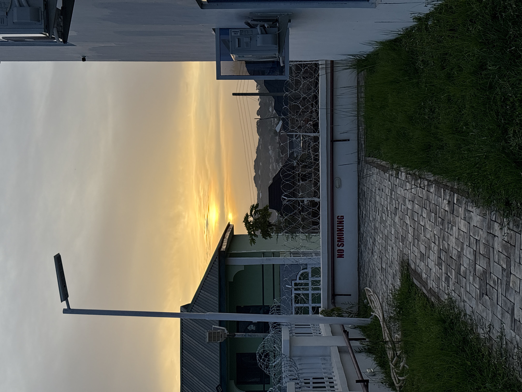 The image captures an outdoor scene at what appears to be a secure compound or property, possibly at sunrise or sunset. The sky, dominating the left side of the frame, displays a striking gradient from a brilliant golden-orange at the bottom, where the sun is a bright disc partially obscured by a roofline, transitioning to a softer pale gray-blue towards the top.
In the foreground, on the right side of the image, there's a paved pathway made of rectangular gray blocks, bordered by lush green grass. A white wall runs alongside this pathway, featuring red, vertically oriented text that reads "NO SMOKING". Further into the scene, a building with a light green facade and a dark, sloped roof is visible, partially enclosed by a white fence topped with intricate razor wire. A small tree stands near this building's roof.
Towards the top-center, another building or structure is seen with a white wall, on which an outdoor utility unit, possibly an air conditioning unit, is mounted. A window or reflective surface below it mirrors the bright sky. A prominent street light with a rectangular fixture extends from the bottom-left corner of the image into the illuminated sky. In the distant background, silhouetted hills or mountains complete the landscape. No people are visible, and there is no discernible activity. The overall lighting suggests a peaceful moment at either dawn or dusk.