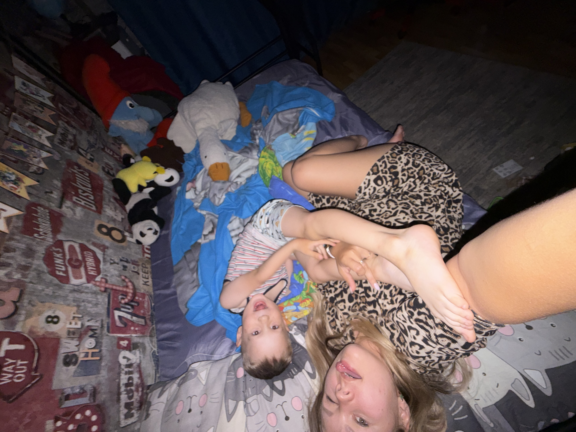 This image captures an intimate moment inside a dimly lit room, likely a bedroom, with the subjects lying playfully on a bed. The photo is rotated 90 degrees clockwise.
**Subjects:** Two individuals are present: an adult female and a young male child. The adult has blonde hair and is wearing a leopard-print top. The child is wearing a striped short-sleeved shirt and patterned shorts. Scattered around them are several stuffed animals, including a prominent white goose, a yellow bear resembling Winnie the Pooh, and a black and white plush toy.
**Objects:** The central object is a bed covered with a combination of blue bedding and grey sheets featuring a repeating cat face pattern. A dark blue curtain is visible in the background, along with a dark rug on the floor.
**Scene/Setting:** The scene is set indoors, characteristic of a casual home environment, specifically a bedroom given the bed and soft toys.
**Activity or event taking place:** Both the adult and the child are relaxed and appear to be engaging playfully with the camera. The adult is winking and sticking her tongue out, while her legs are raised, with one foot visible near her face. The child has an open-mouthed expression, suggesting laughter or excitement.
**Notable details:** The low light levels suggest it might be nighttime or a poorly lit room, contributing to a cozy, relaxed atmosphere. There are no overt weather cues. The emotions conveyed are joyful, playful, and carefree.
**Visible text:** On the wall to the left (top in the rotated view), there are decorative signs and posters with various text: "FUNK'S", "HYBRID", "SWEET HOME", "WAY OUT", "7 Up", "Mobil 1", "Biskuits", "Schokolade", and the number "8" twice. An anchor symbol is also visible.