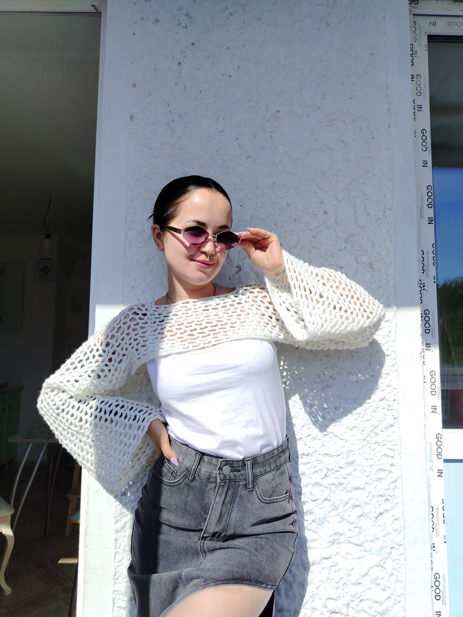 Here is a concise description of the image:
A young woman stands outdoors, likely on a balcony or near an entrance, judging by the presence of a door frame or window frame. The text "GOOD IN" is repeatedly printed on a white frame, which could belong to a window or door. The woman is wearing a white top and denim skirt, with a cropped crocheted white sweater. She's holding a pair of pink-tinted sunglasses and looking slightly to the side. The lighting suggests it is a sunny day. The location details (Noviy, Russia) are not directly discernible from the image itself.