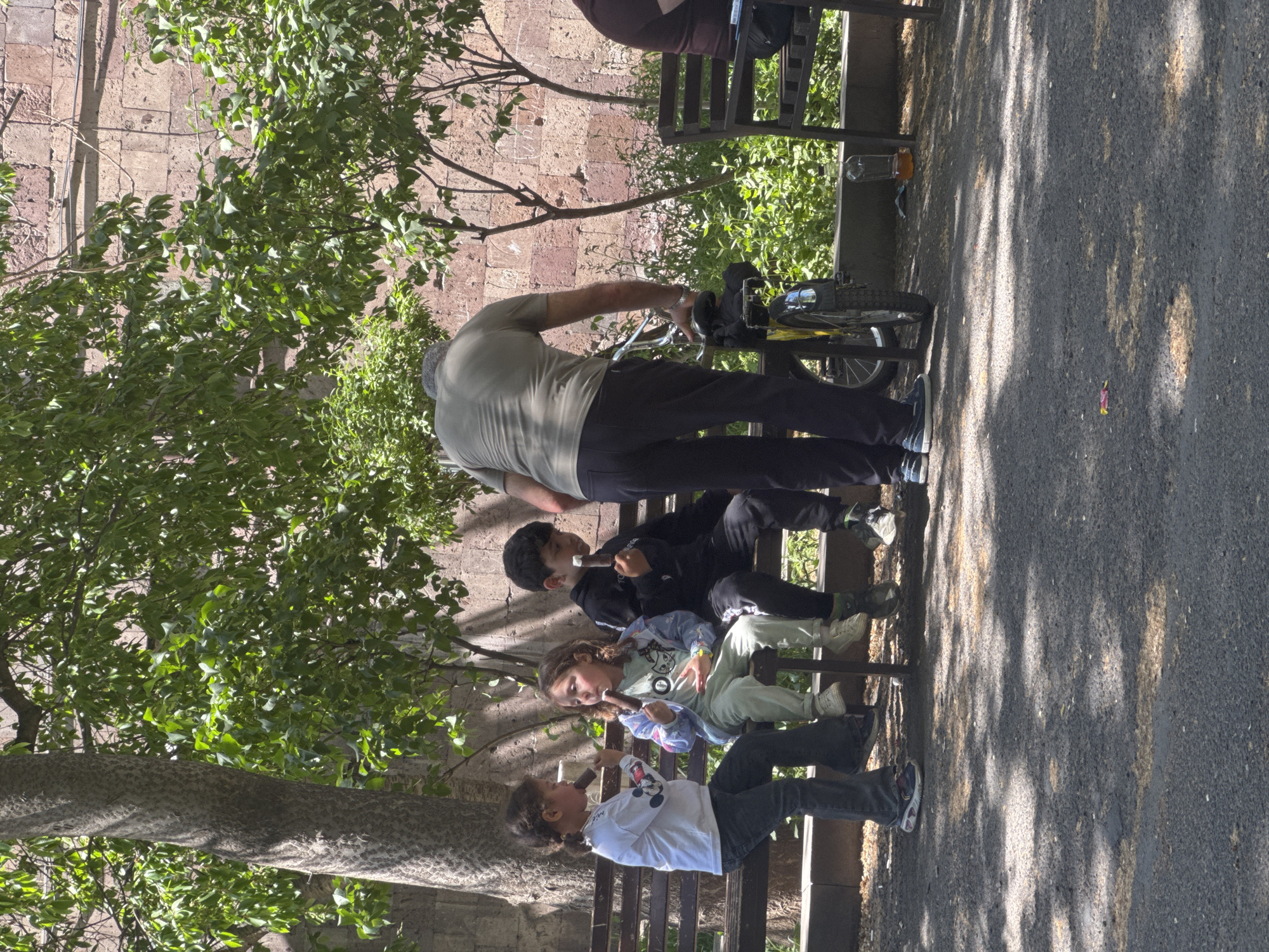 The photograph captures a scene in Yerevan, Armenia. Three children sit on a park bench, each eating an ice cream bar. An adult stands nearby, appearing to tend to a bicycle. The setting includes a paved walkway, a stone wall, and trees, suggesting a park or green space within the city.